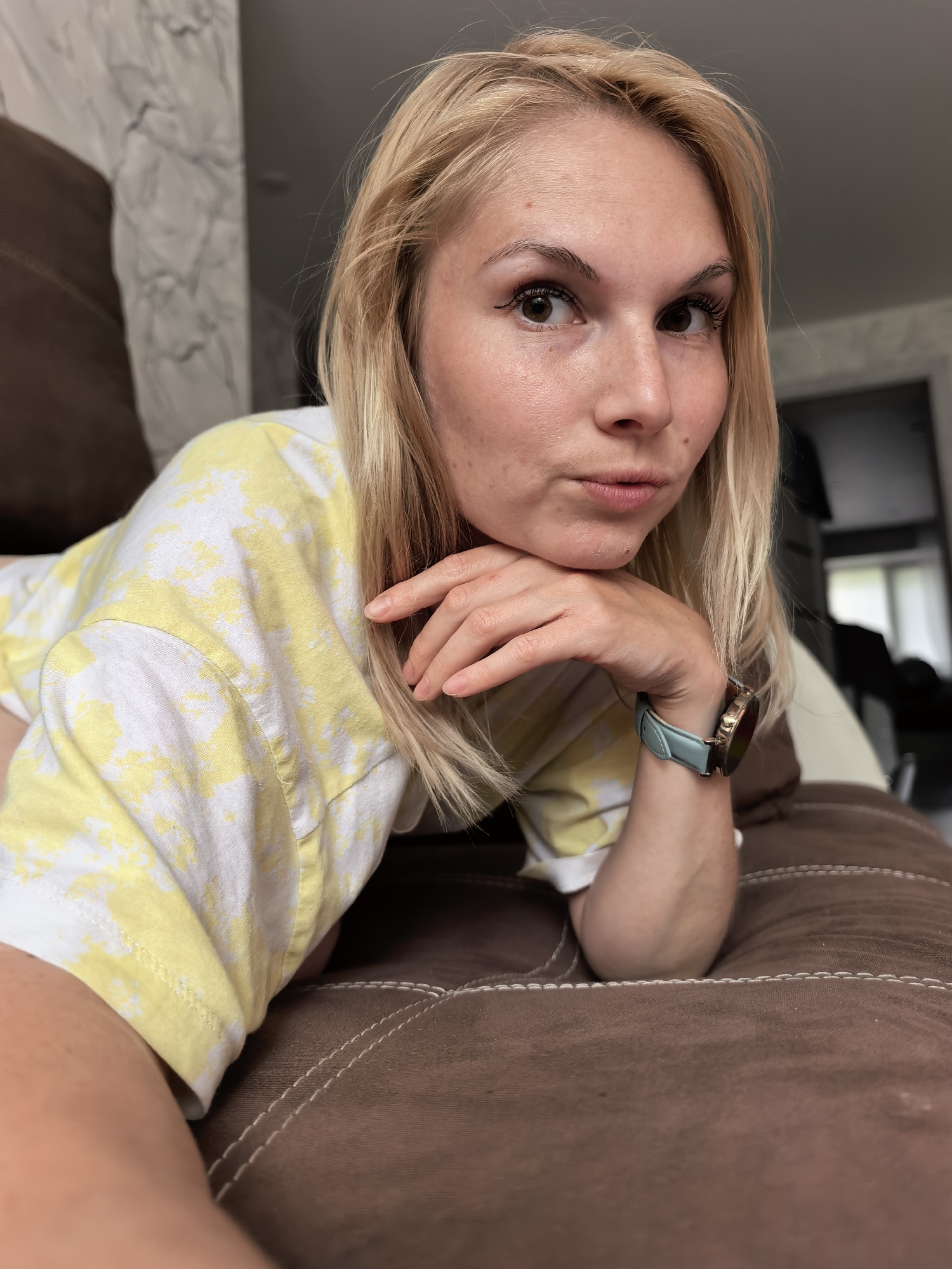 The image, identified as taking place in Sterlitamak, Russia, depicts an adult blonde female positioned prone on a brown, upholstered surface, likely a sofa or bed, characterized by white decorative stitching. Her left arm is bent, and her hand supports her head, resting under her chin and jawline. She wears a loose-fitting white t-shirt with a pale yellow tie-dye or mottled pattern. A light blue/teal watch with a circular, gold-toned face is visible on her left wrist. Her gaze is directed straight at the viewer. Her blonde hair is casually styled around her face. She has brown eyes with visible eyeliner and mascara, a fair complexion, and minor blemishes are visible on her forehead and chin. The background consists of an indoor room environment, with a light-toned, possibly marble-patterned wall segment visible to the left, and a blurred, undifferentiated room interior to the right. The lighting is soft and even. The image offers no specific visual identifiers of Sterlitamak, Russia.