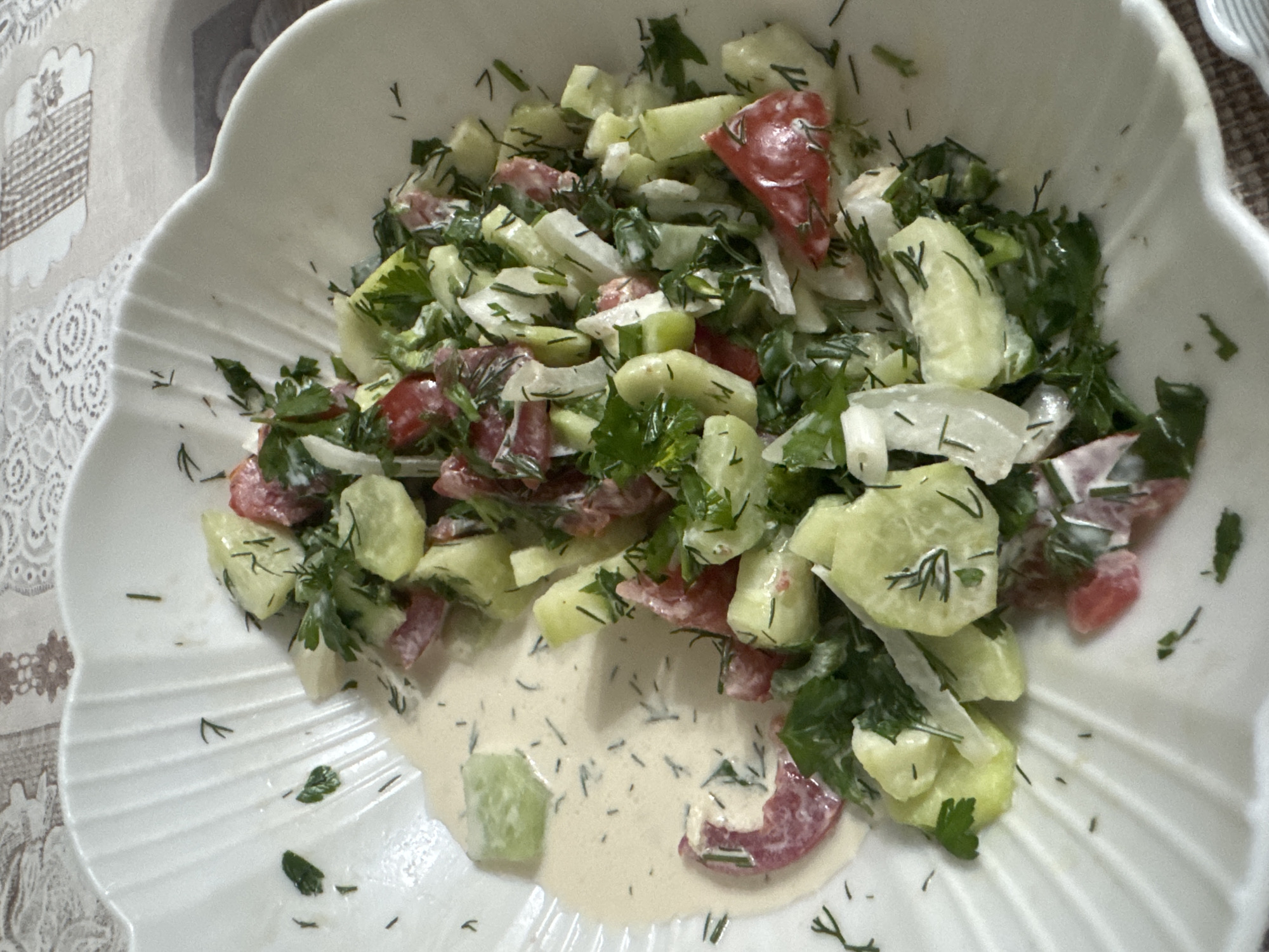 A close-up shot captures a white, fluted bowl filled with a fresh salad. The salad is composed of chopped cucumbers, tomatoes, and onions, all tossed in a creamy white dressing and garnished with fresh dill and parsley. A single slice of cucumber sits apart from the main salad in a pool of the dressing on the left side of the bowl. The bowl rests on a patterned tablecloth. The image appears to be taken indoors, likely in a dining setting in Таганрогский, Russia.