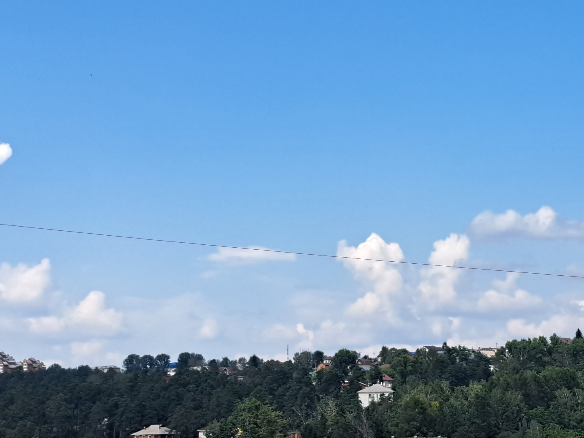The image depicts a panoramic view of a hillside settlement under a bright blue sky with scattered cumulus clouds. The foreground is dominated by a dense, dark green forest, suggesting a natural or semi-natural landscape. Several houses are visible nestled within and on top of the treeline, indicating a residential area. The architecture of the houses appears to be varied, with some featuring pitched roofs and light-colored facades. A thin, dark wire stretches horizontally across the upper portion of the image, possibly a power line or telecommunication cable. The overall scene is one of a peaceful, somewhat remote, or suburban environment. Without specific landmarks, it is impossible to definitively pinpoint the location within Irkutsk, Russia, but the presence of residential buildings atop a forested slope is characteristic of many areas around the city, especially those bordering the taiga.