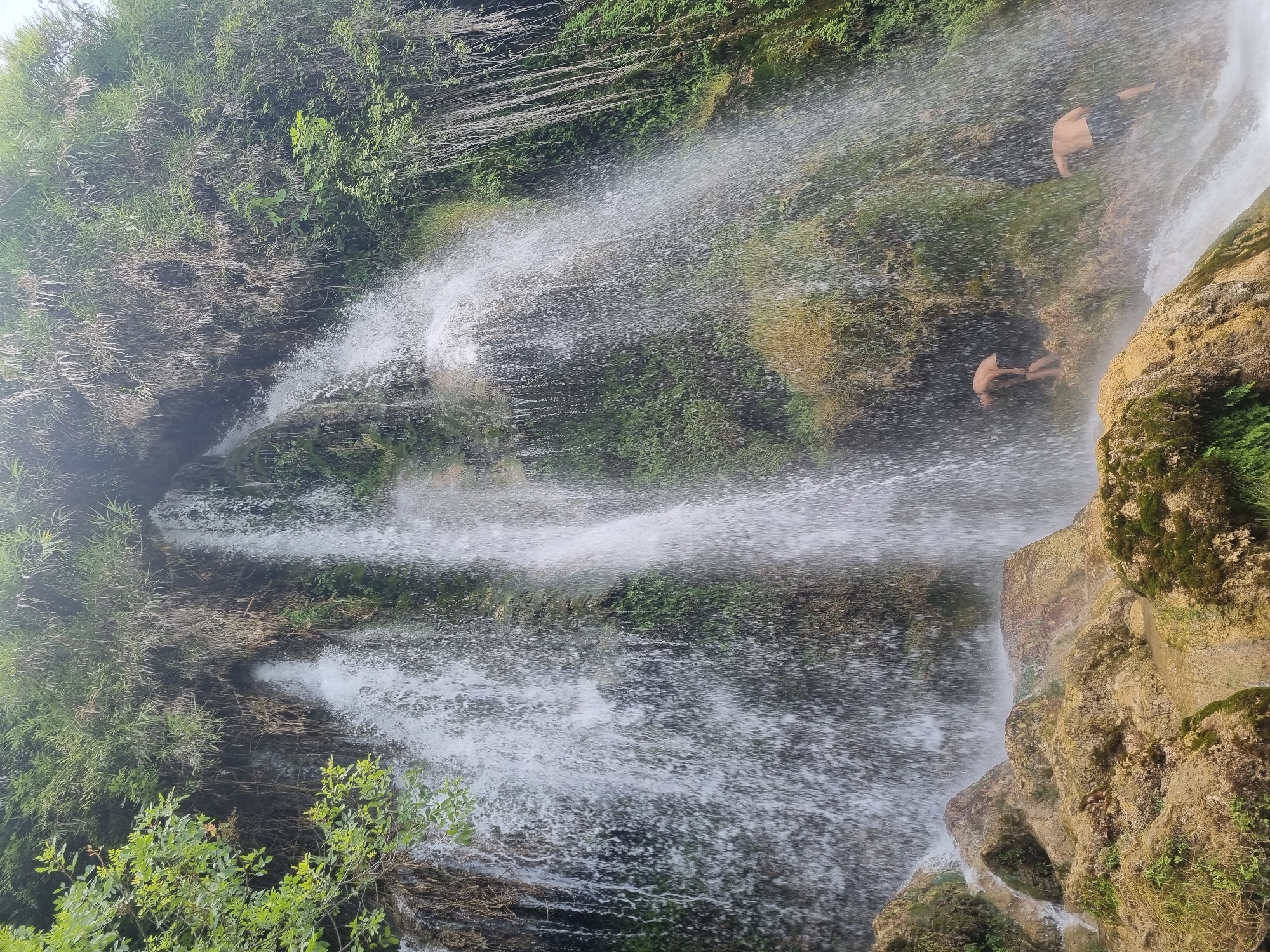 A wide shot captures a cascading waterfall at the base of a lush, green cliff face. Two young men, one in the foreground and one slightly behind him, are positioned near the base of the waterfall, partially obscured by the spray. The man in the foreground, facing away from the camera and to the right, appears to be wearing dark shorts. The second man, to the left, is also facing away from the camera and is bent over. The waterfall itself is dense and powerful, with water falling in many streams and sheets, creating a misty environment. The cliff is covered in green vegetation and moss, with some rocky outcrops at the bottom. The overall scene suggests a natural, outdoor location, possibly a popular spot for swimming or recreation. The location of this waterfall is in Anna, Spain.