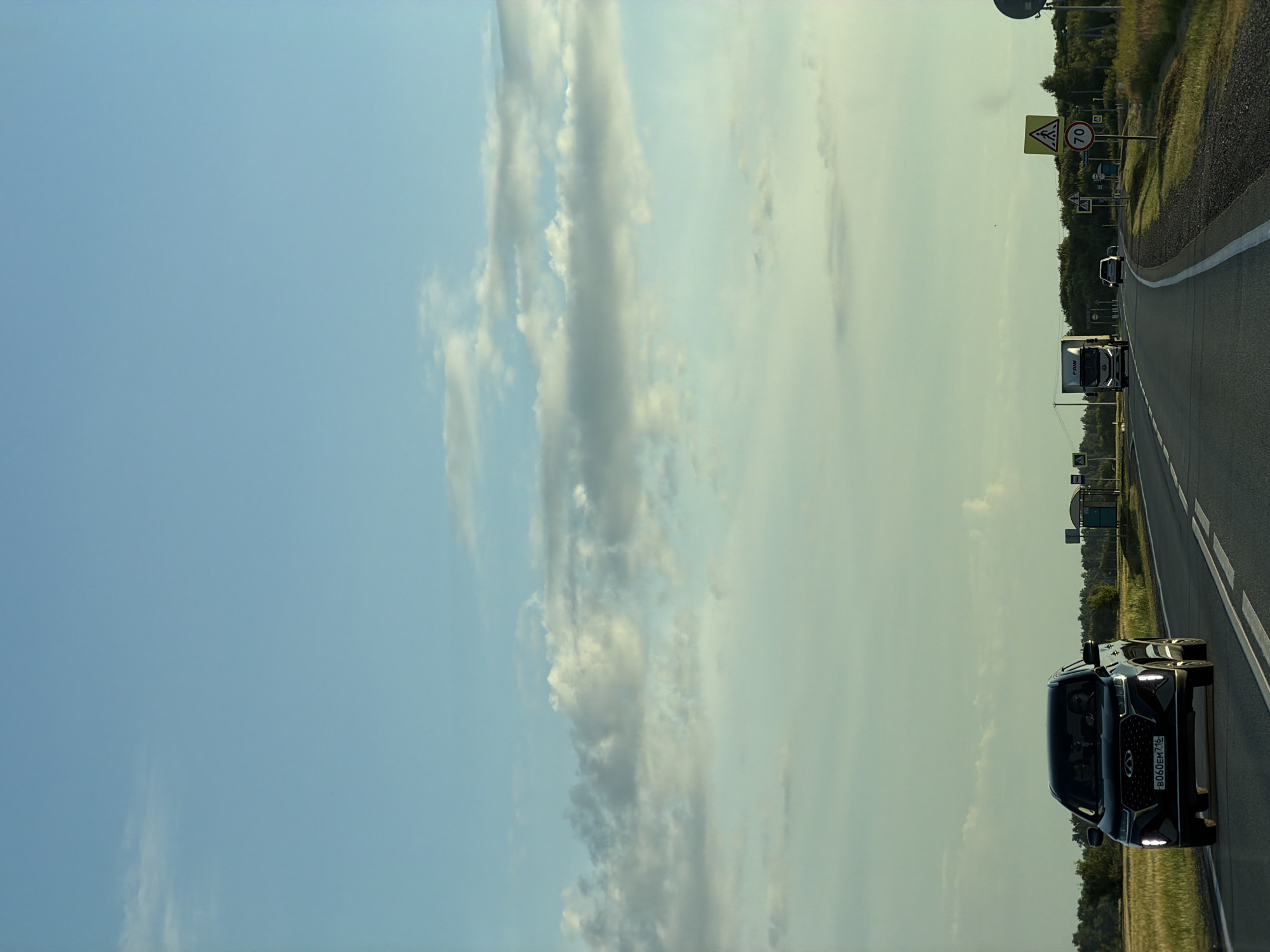 An exterior, eye-level, medium shot captures a road in Aryshkhazda, Russia, under a clear sky with scattered clouds. In the foreground, a black SUV is driving on the right side of a two-lane asphalt road. The SUV has its headlights on, and a license plate reading "B060EM 716" is visible. Further down the road, a white truck and a white car are also traveling. On the right side of the road, there are several road signs, including a pedestrian crossing sign and a speed limit sign indicating 70. The landscape surrounding the road consists of fields and trees.