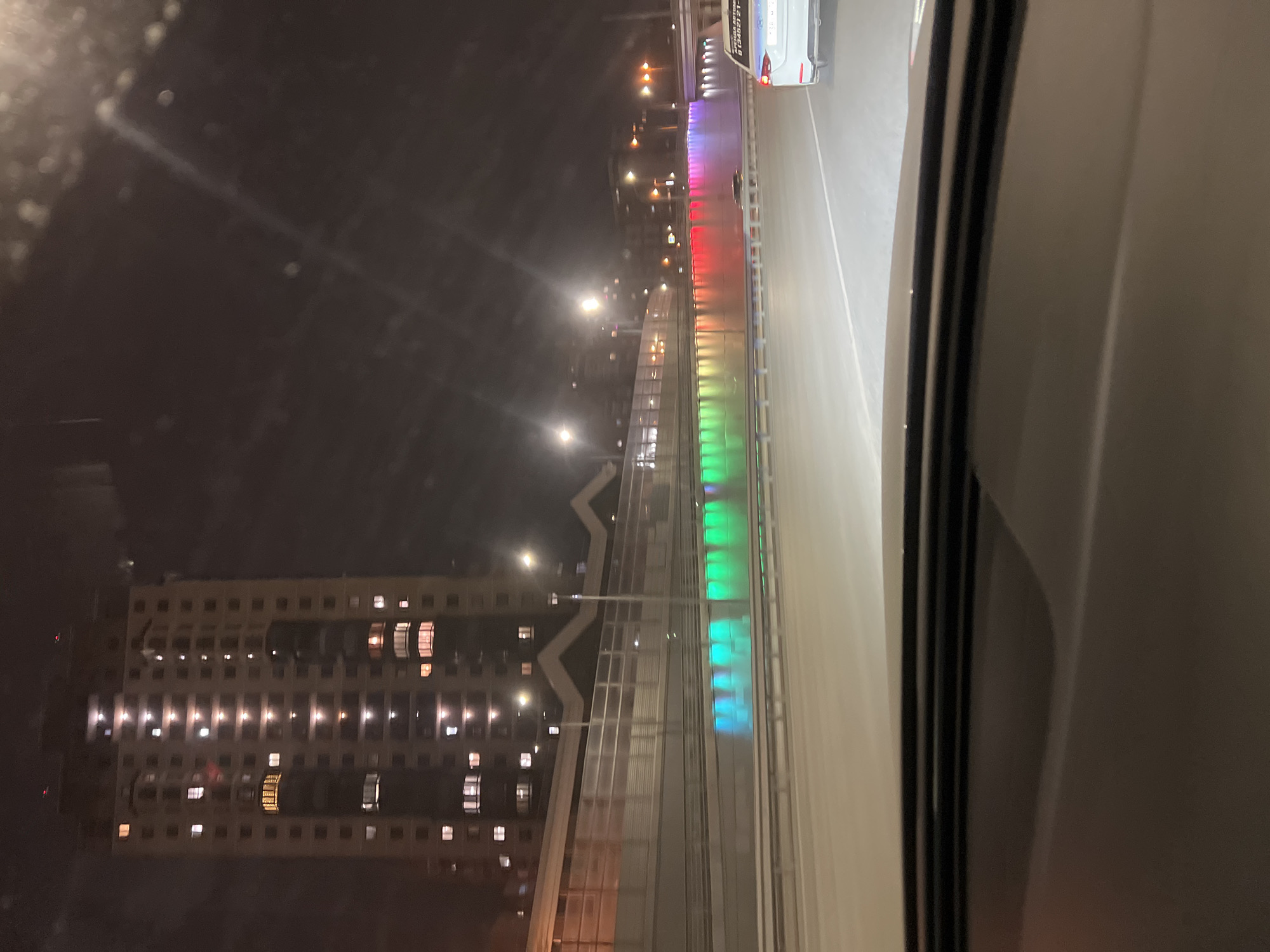 The image shows an elevated roadway at night, viewed from inside a building through what appears to be a window. The roadway is illuminated with colored lights that transition from green to red, indicating a possible pedestrian or cycle path. A building with many illuminated windows is visible in the background. The scene is located in the city of Tyumen, Russia. A vehicle is partially visible on the roadway, and the overall setting suggests an urban environment.