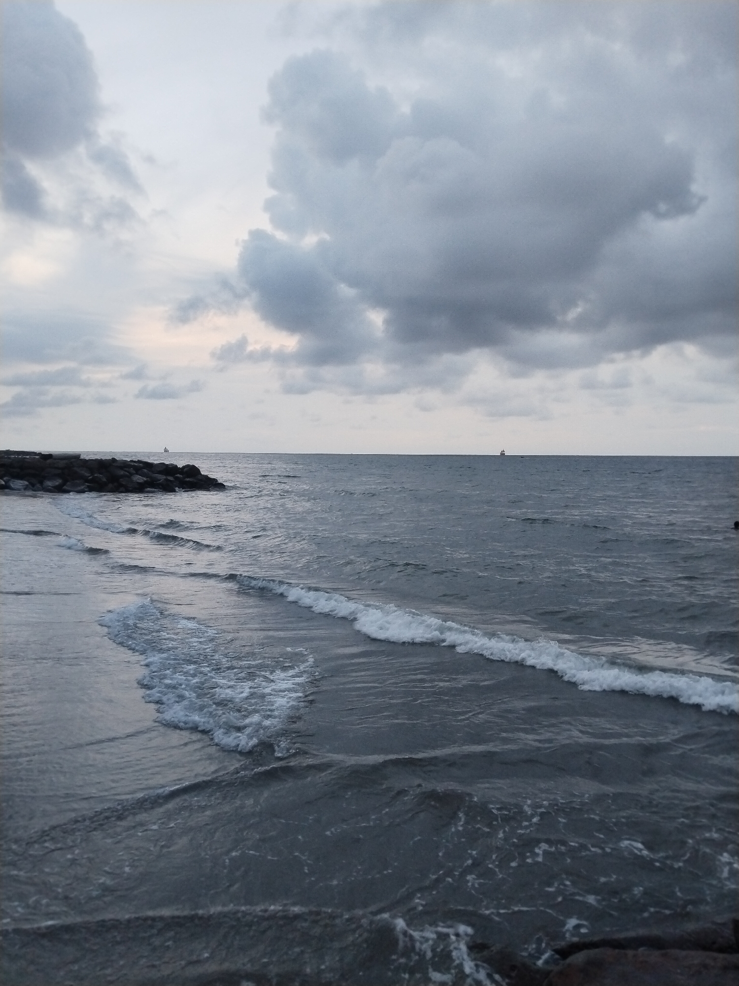 The image shows a seascape under a cloudy sky, likely at dusk or dawn due to the muted lighting. In the foreground, the shoreline is visible with dark rocks and sand meeting the ocean. Waves are breaking along the shore, creating white foam against the dark water. In the distance, a small boat or ship can be seen on the horizon. Given the prompt, the location is inferred to be in Lhokseumawe, Indonesia. There are no people visible in the image. The overall scene conveys a sense of calm and overcast weather.