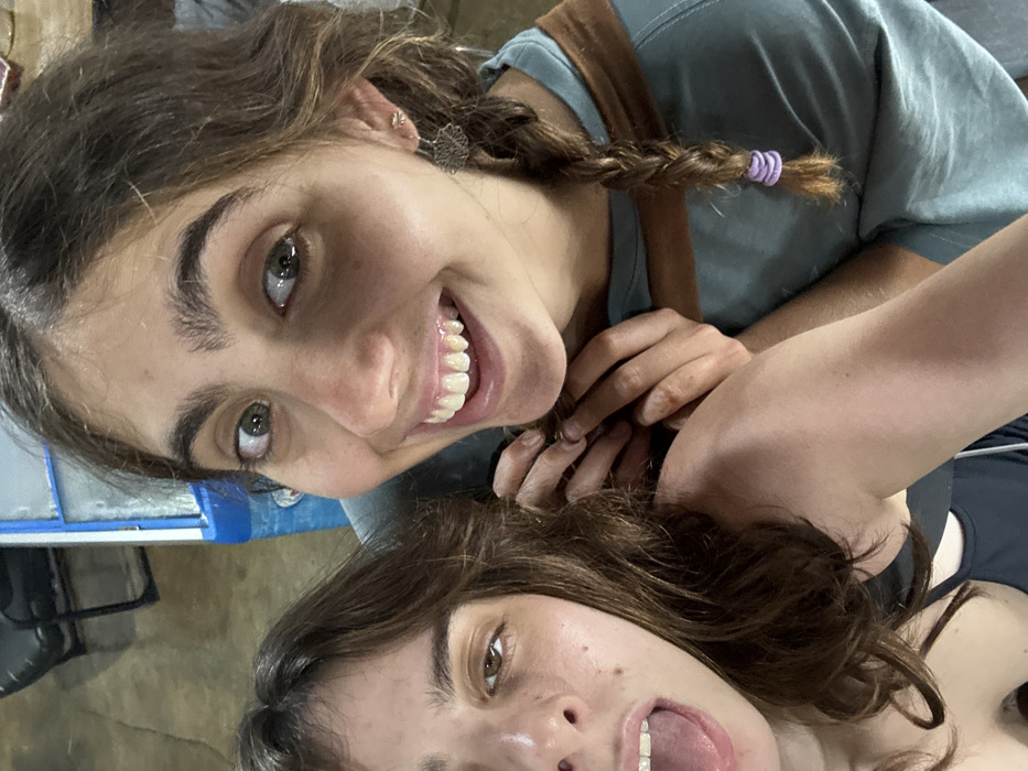 Two young females are depicted in a close-up, informal photograph. The prominent female, positioned right of center, has dark brown hair, which includes a braid secured with a purple elastic on the visible side. She has prominent eyebrows, green eyes, and a broad smile revealing upper teeth. She wears a light teal shirt with a brown accent and two small, intricate silver-toned earrings. Her hands are clasped near the shoulder of the second female. The second female, partially visible on the left, has dark brown wavy hair, her mouth open with her tongue slightly extended, and visible dental braces on her upper teeth. She wears a dark-colored top. The background is out of focus, showing indistinct elements including a light brown surface, a dark object, and a segment of a blue and white structure, consistent with an indoor or covered outdoor setting. Lighting appears artificial. The image contains no identifiable landmarks or specific features to determine a precise location within Thap Sakae, Thailand.