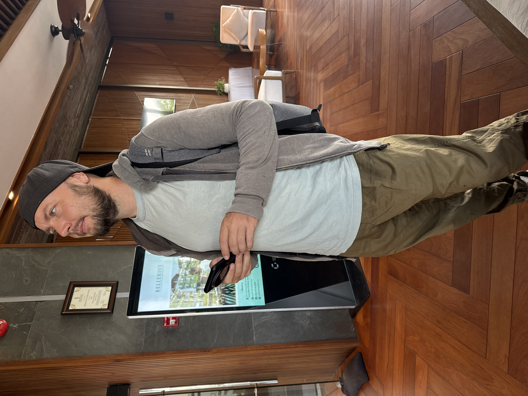 The image depicts a man standing indoors, likely at a residential property development in Đà Nẵng, Vietnam, judging by the "BELLERIVE" signage on the nearby interactive display. The man is wearing a gray hat, light gray t-shirt, gray hooded jacket, and olive green pants. He is holding a black smartphone or similar device. The location is characterized by wooden flooring, paneled walls, and modern furnishings including white chairs and cushions. An award certificate is mounted on the wall next to the interactive display. The overall ambiance suggests a high-end or luxury setting.