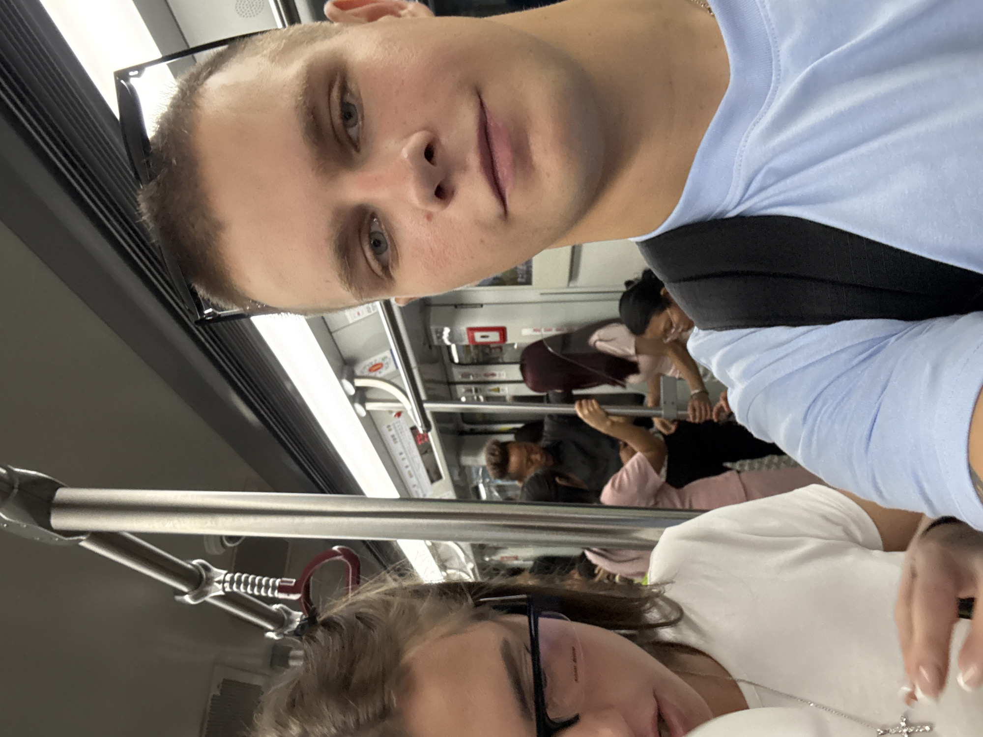 In Haizhu District, China, a man and woman are seated on a train. The man, in the foreground, is taking a selfie with the woman beside him. He has short brown hair and is wearing a light blue t-shirt and a black backpack. The woman is wearing glasses and a white t-shirt, with a silver cross necklace. Several other passengers are visible on the train, some standing and holding onto the overhead rails. The train carriage appears to be brightly lit with fluorescent lights.