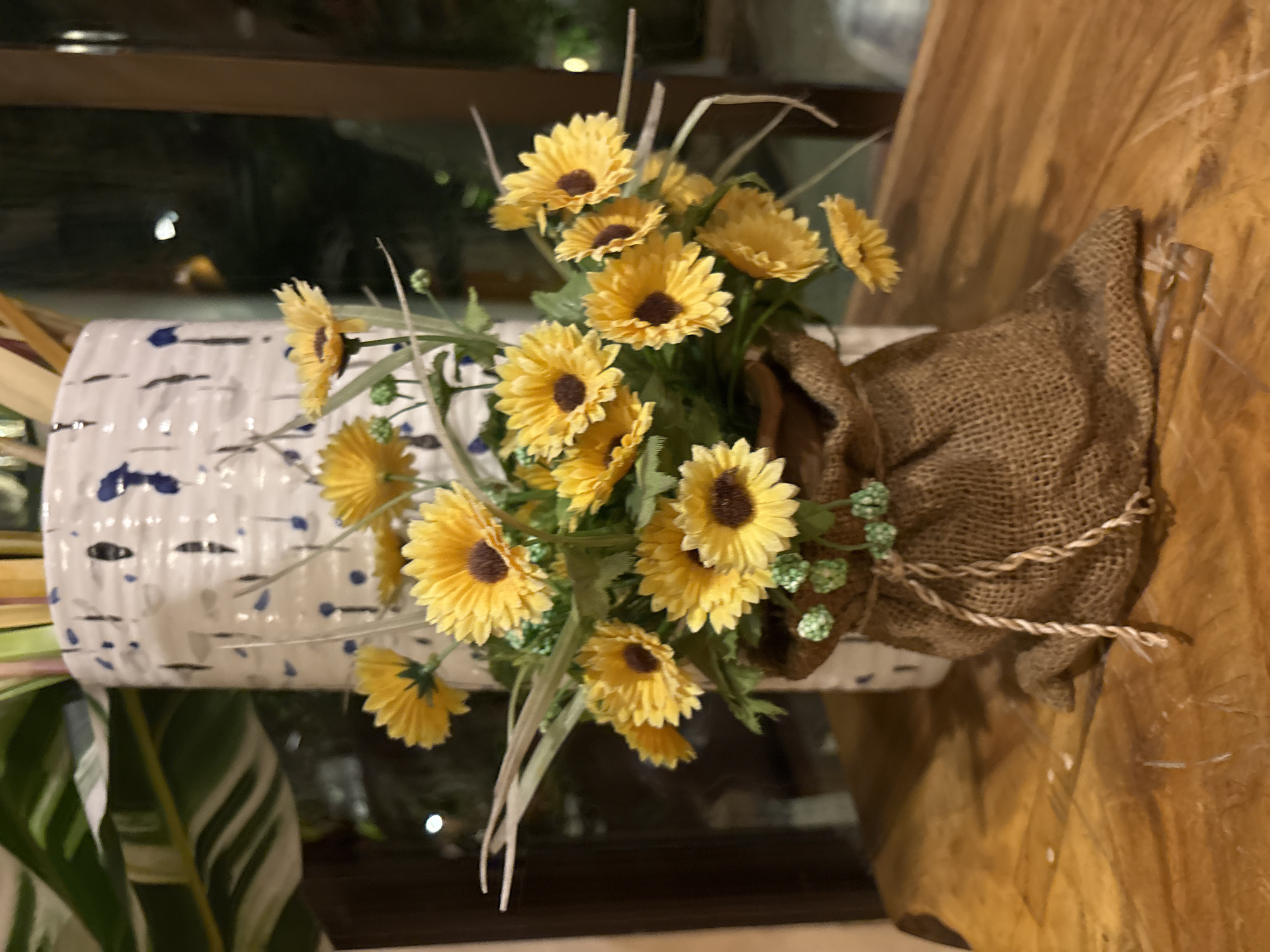 A vase of artificial yellow sunflowers sits on a wooden table. The vase is white with blue splatters and is partially covered by a burlap sack tied with rope. In the background, a large leafy green plant is visible, along with a window showing a dark, possibly night-time, outdoor scene. There are no people or animals present. The setting appears to be indoors, possibly a restaurant or home, in Chalong, Thailand.