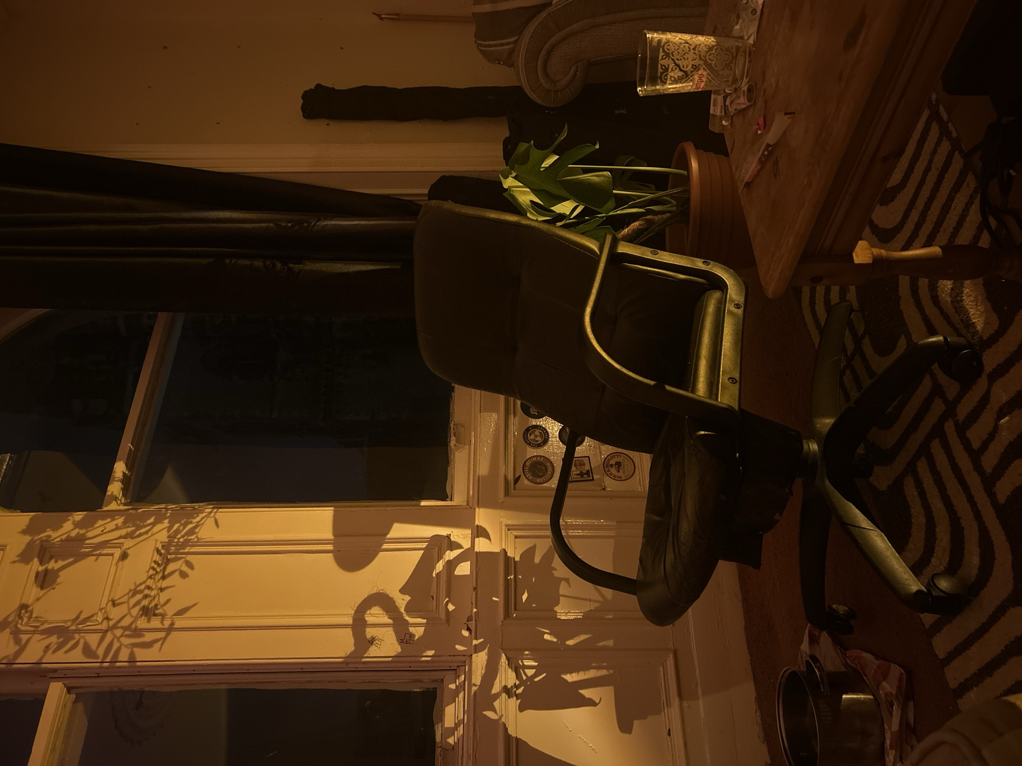 The image captures an indoor scene, likely a room within a residential setting in Dundee, United Kingdom, characterized by dim, warm lighting that suggests evening or nighttime. The scene is rotated 90 degrees clockwise.
In the correct orientation, a dark, wheeled office chair occupies the central foreground. Behind it, a large houseplant, possibly a Monstera, stands in a terracotta pot. To the chair's right, a wooden table holds a clear glass with an ornate dark pattern and a few other indistinct items. Below the table and chair, a rug features a bold striped or zigzag pattern in dark and light tones.
To the left, a window area is partially covered by dark curtains. Striking shadows of plant foliage are cast across the light-colored window frame and the wall beneath, indicating a strong light source, possibly from outside or positioned to create these effects. The light-colored wall in the background shows a section of a sofa or armchair with decorative carvings. Several small stickers or badges are adhered to the wall near the window frame. On the floor near the chair's base, a couple of metallic pots or bowls are partially visible.
No specific activities or people are present; the image is a still life, emphasizing the interplay of light and shadow within the room. While there is text on the patterned glass and the wall stickers, it is not legible.