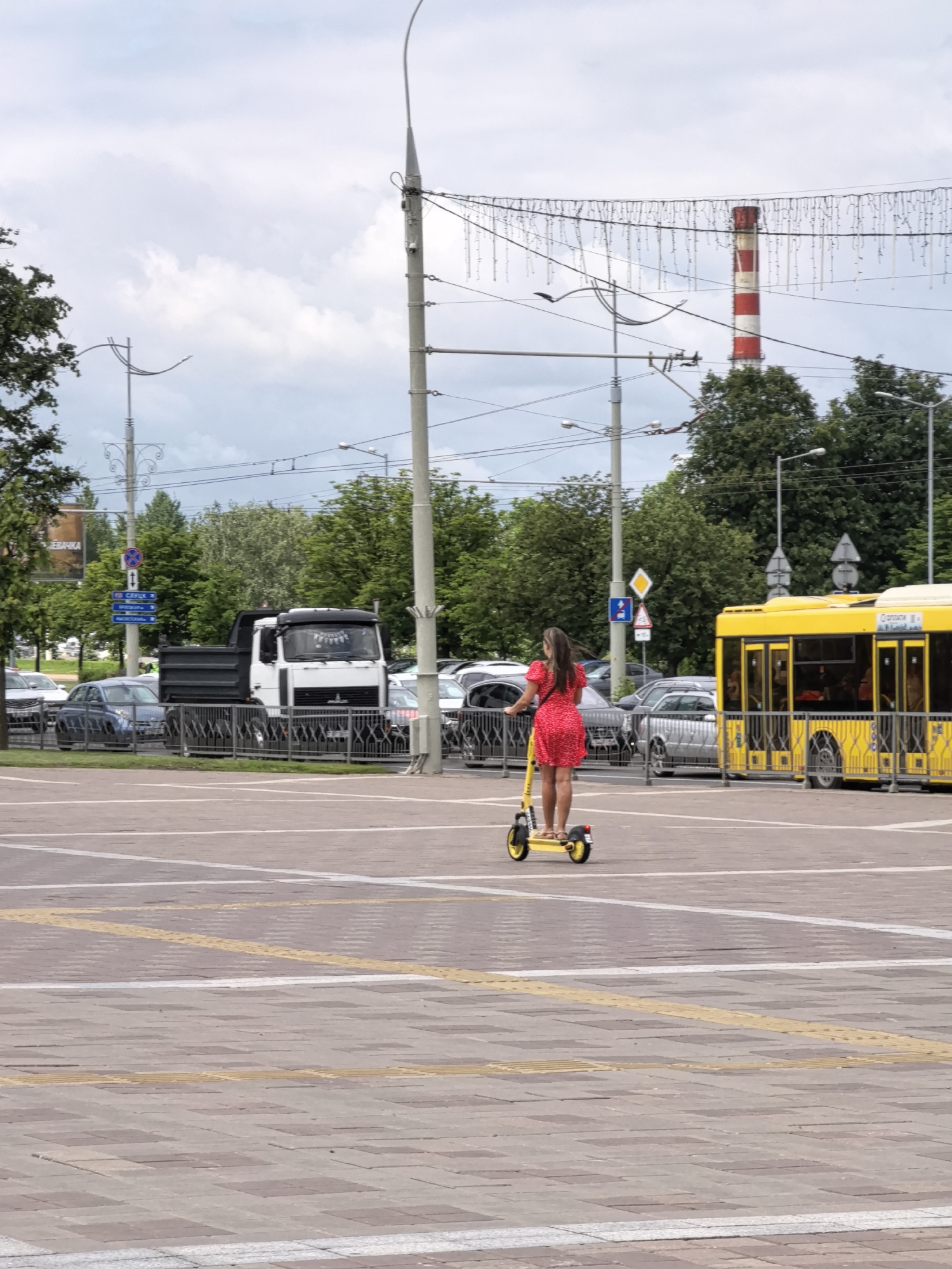 In the city of Unknown, Unknown, a woman with long brown hair wears a short, red, floral-print dress and tan sandals as she rides a yellow electric scooter on a paved public square. The scooter's handlebars and wheels are also yellow. Behind her, a bright yellow public bus with tinted windows is parked. Further back, a white dump truck with a dark gray bed is stopped on a street, bordered by a metal fence. Several cars are visible in the background, along with lush green trees and a tall industrial chimney with red and white stripes. Streetlights and overhead wires crisscross the sky, which is partly cloudy. The ground is paved with square tiles, with yellow tactile paving lines indicating a pedestrian crossing.