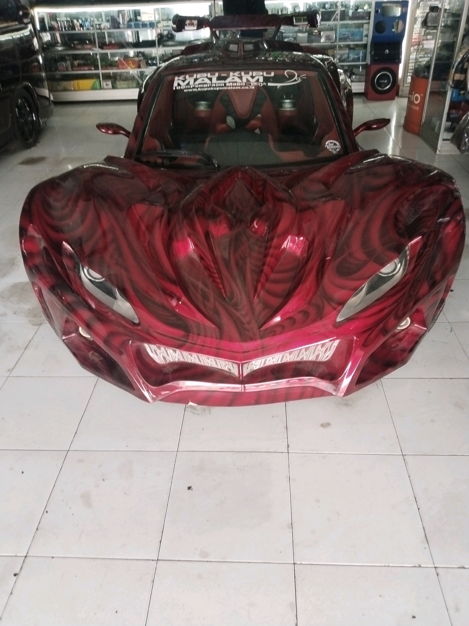 A custom red sports car with flame-like black airbrushed accents is parked indoors on a tiled floor. The car is positioned head-on, showcasing its aggressive front design, sharp headlights, and a wide grille. The interior appears to be a custom build with red and black upholstery. In the background, shelves are stocked with various items, suggesting this might be a retail or workshop environment. The car's custom paint job and aggressive styling are the focal points of the image. The environment appears to be a commercial space located in Sinduadi, Indonesia.