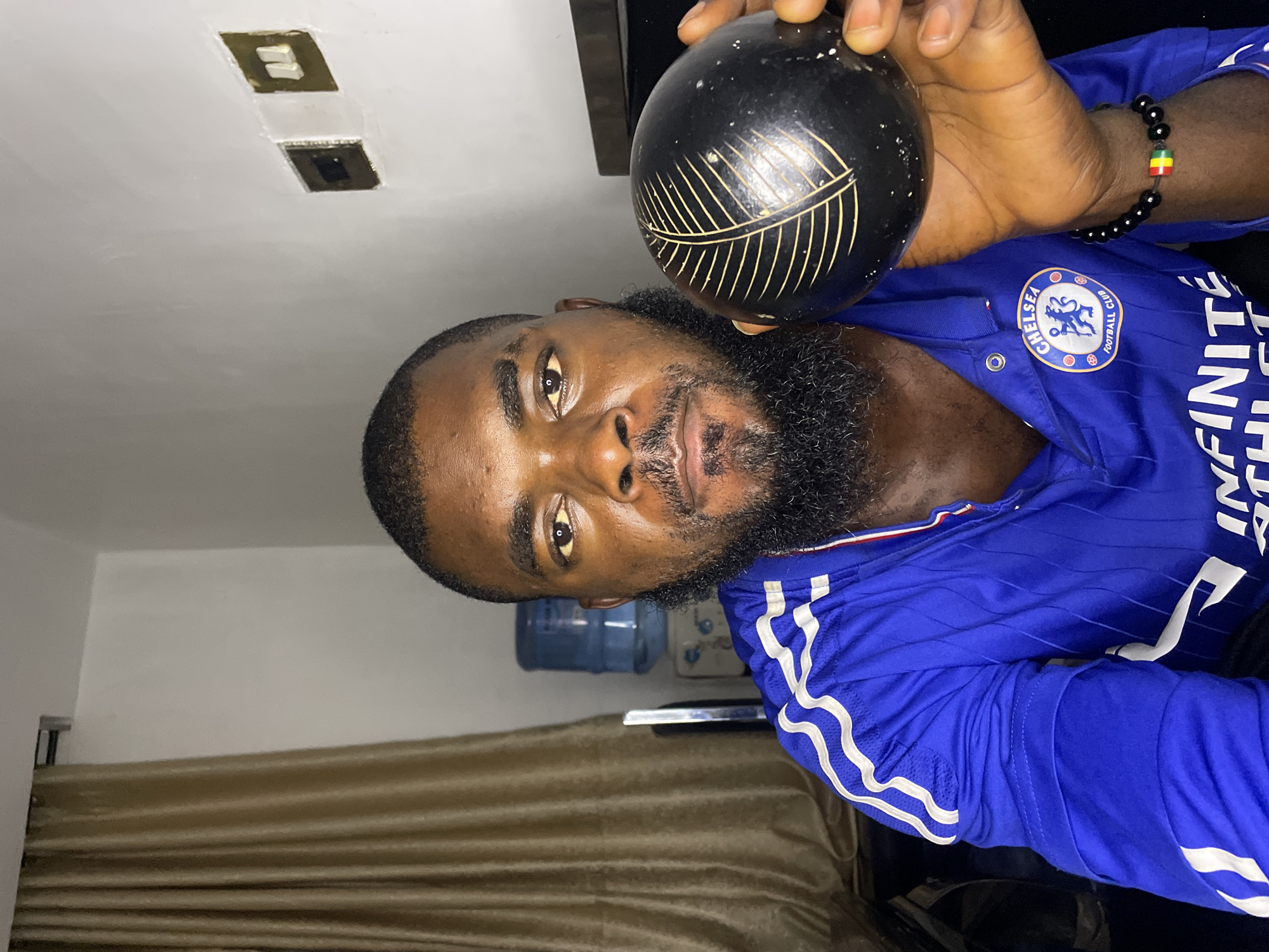 A medium shot captures a man in a blue Chelsea Football Club jersey holding a black, spherical object with a leaf-like design. He has a beard and is looking directly at the camera. Behind him, a beige curtain hangs on the left, and on the right, a white wall features two electrical outlets, one of which appears to be damaged. A blue water dispenser is visible in the background. The scene appears to be indoors.