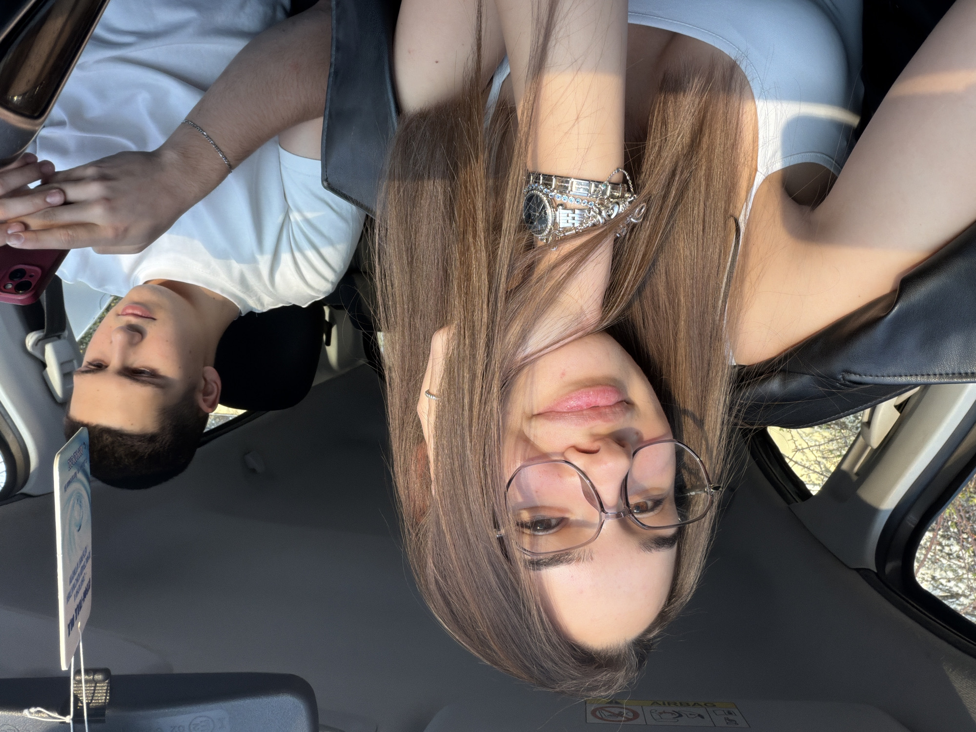 A selfie of a young woman with long brown hair and glasses sitting in a car with a young man. The woman is in the passenger seat wearing a white tank top, and a silver watch on her left wrist. The man is in the driver's seat, wearing a white t-shirt, holding a red phone. The interior of the car is dark, with a car freshener hanging from the rearview mirror. The car is parked, and trees can be seen through the car windows. Location is unknown and cannot be determined from this image.