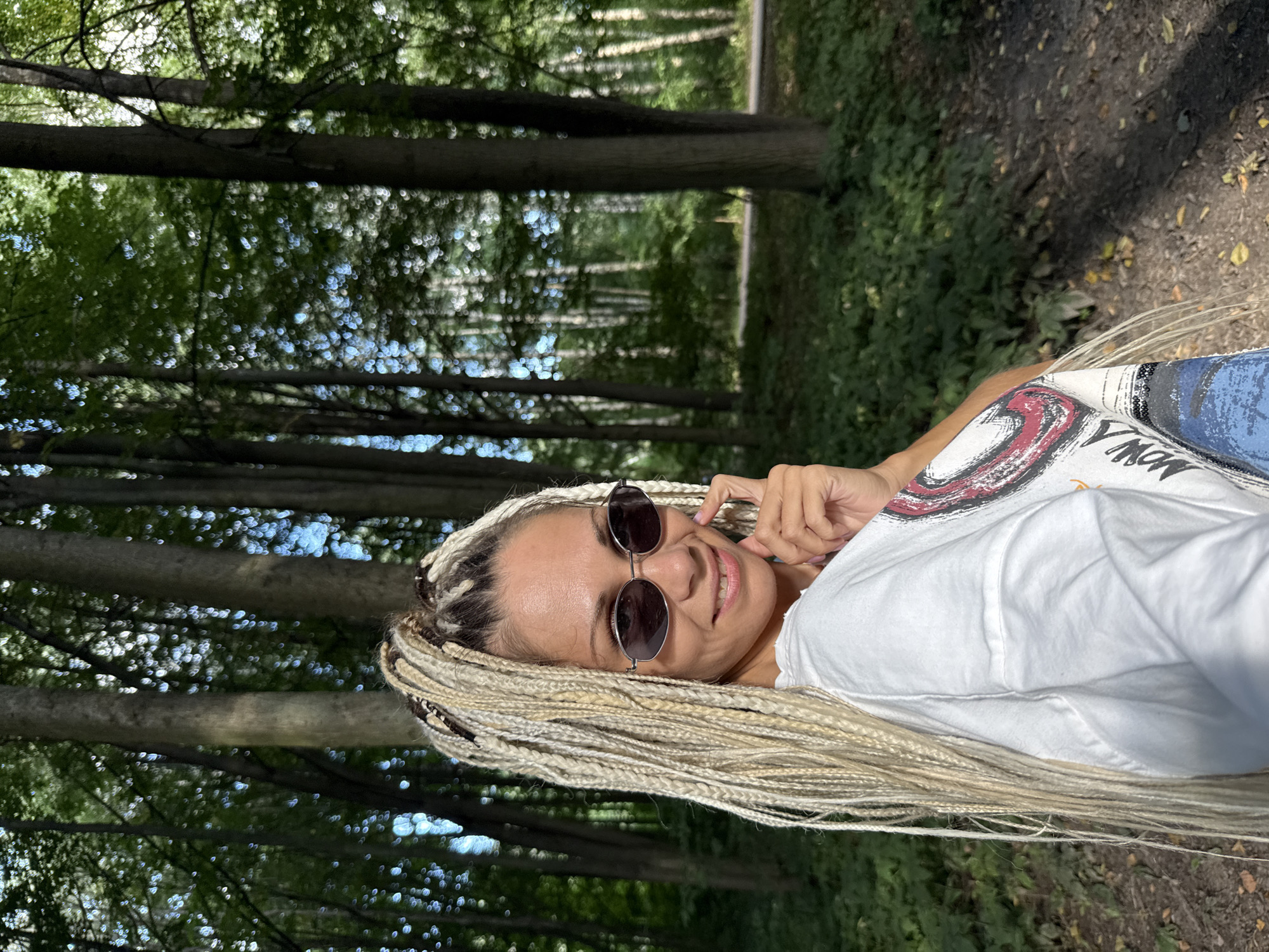 A woman with blonde braids stands in a forest in Rusanovo, Russia, facing the camera and smiling. She is wearing sunglasses and a white t-shirt with a graphic print. The background is filled with tall trees and dappled sunlight.