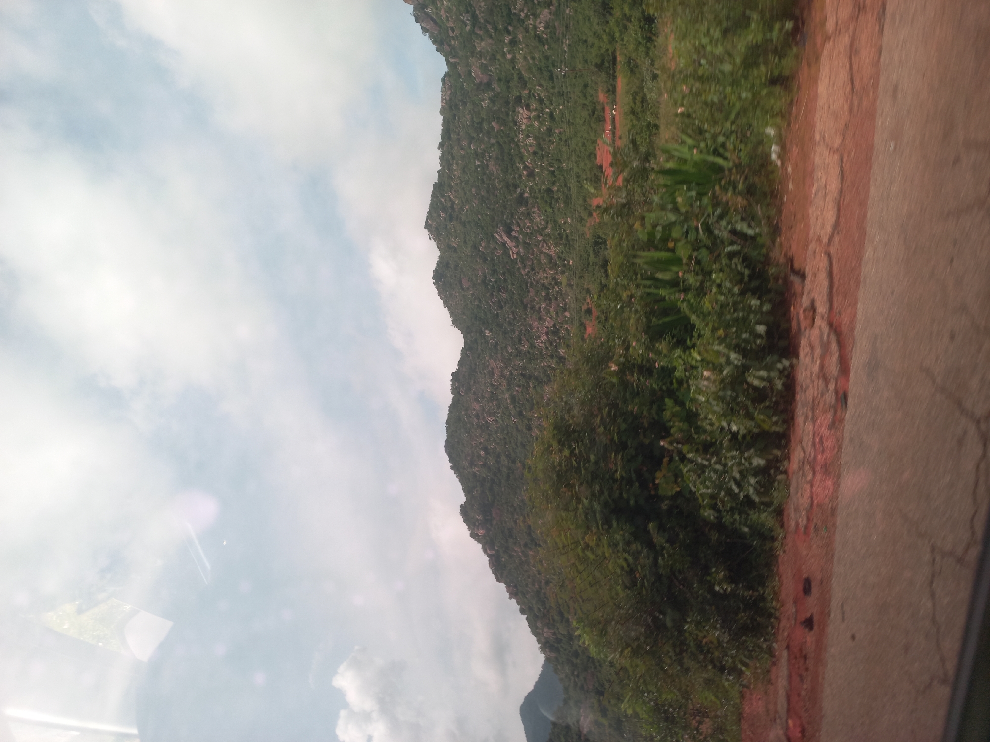 A wide shot captures a mountainous landscape with dense vegetation and a cloudy sky. The foreground features a cracked asphalt road and reddish-brown soil. In the midground, lush green trees and bushes line the base of a rocky mountain covered in thick foliage. The mountain itself has a rugged, uneven terrain with scattered large rocks. The sky is partially obscured by white and gray clouds, with hints of blue peeking through. The overall scene is natural and undeveloped, suggesting a rural or wilderness setting.