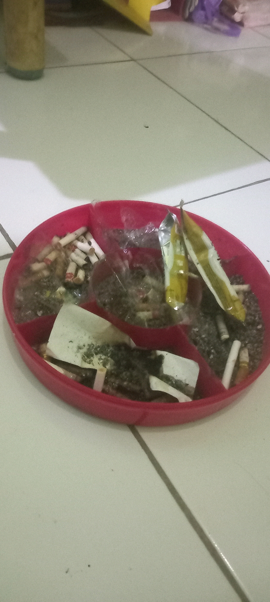 The image shows a red, segmented plastic tray filled with cigarette butts, ash, and what appears to be marijuana. There are at least two partially smoked marijuana cigarettes with yellow wrappers. The tray is on a light-colored tiled floor, possibly in a residential setting. The lighting is dim, suggesting an indoor environment. There is no indication of the location being specifically in Argasoka, Indonesia.