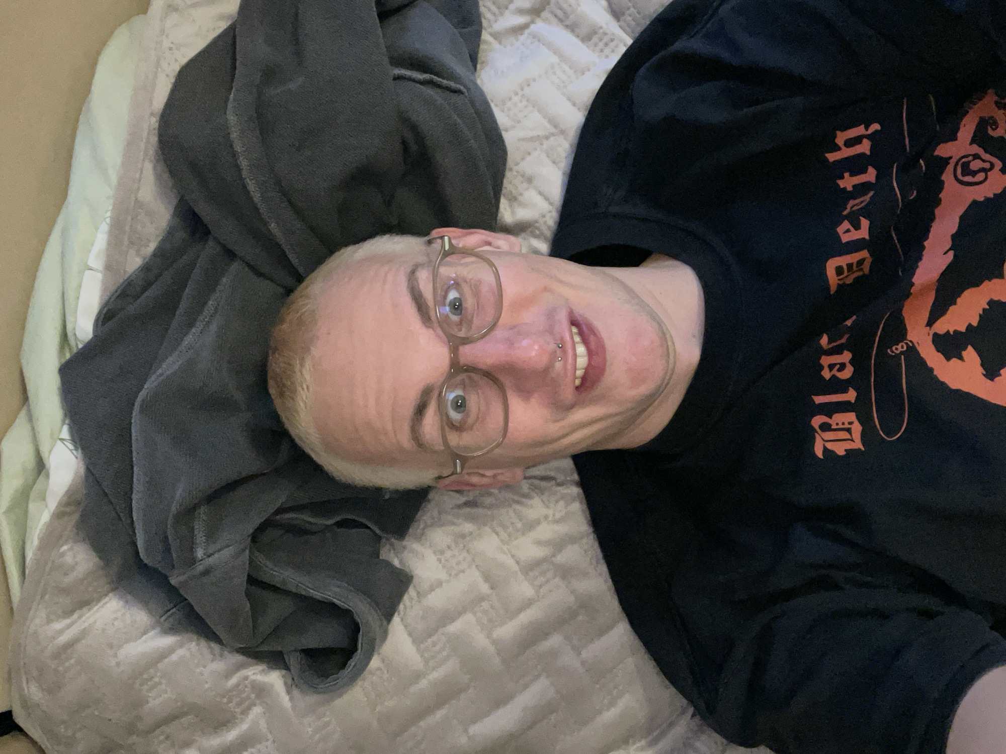 A person with short, dyed blonde hair and round glasses lies on a bed. They are wearing a black sweatshirt with orange lettering that appears to say "Black Death." The person has a septum piercing. The bed is covered with a quilted comforter. Dark grey fabric is draped over the top of the frame. The scene appears to be indoors, likely a bedroom. There is no indication of the city of Rozhok, Russia, in the image.