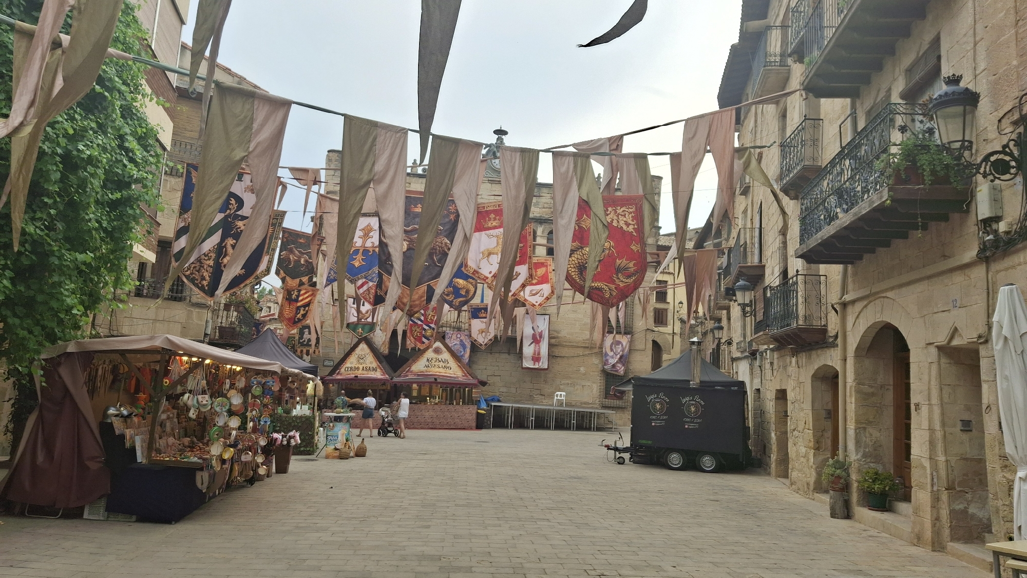 The image depicts a street scene in La Fresneda, Spain, likely during a medieval-themed event. Numerous banners with heraldic crests and historical motifs are strung across the street between buildings. On the left, a market stall displays various handicrafts and trinkets under a brown awning. Further into the scene, two food stalls are visible: one labeled "CERDO ASADO" (Roast Pork) and another "KEBAB ARTESANO" (Artisan Kebab). A black food truck, branded "LUIGI'S PIZZA," is parked on the right side of the street. Several people are present, some browsing the stalls, while others appear to be staff. The architecture of the surrounding buildings features stone facades and balconies with wrought-iron railings, indicative of traditional Spanish urban design. The cobblestone street is clean and well-maintained. The sky is overcast, suggesting potentially cooler weather.