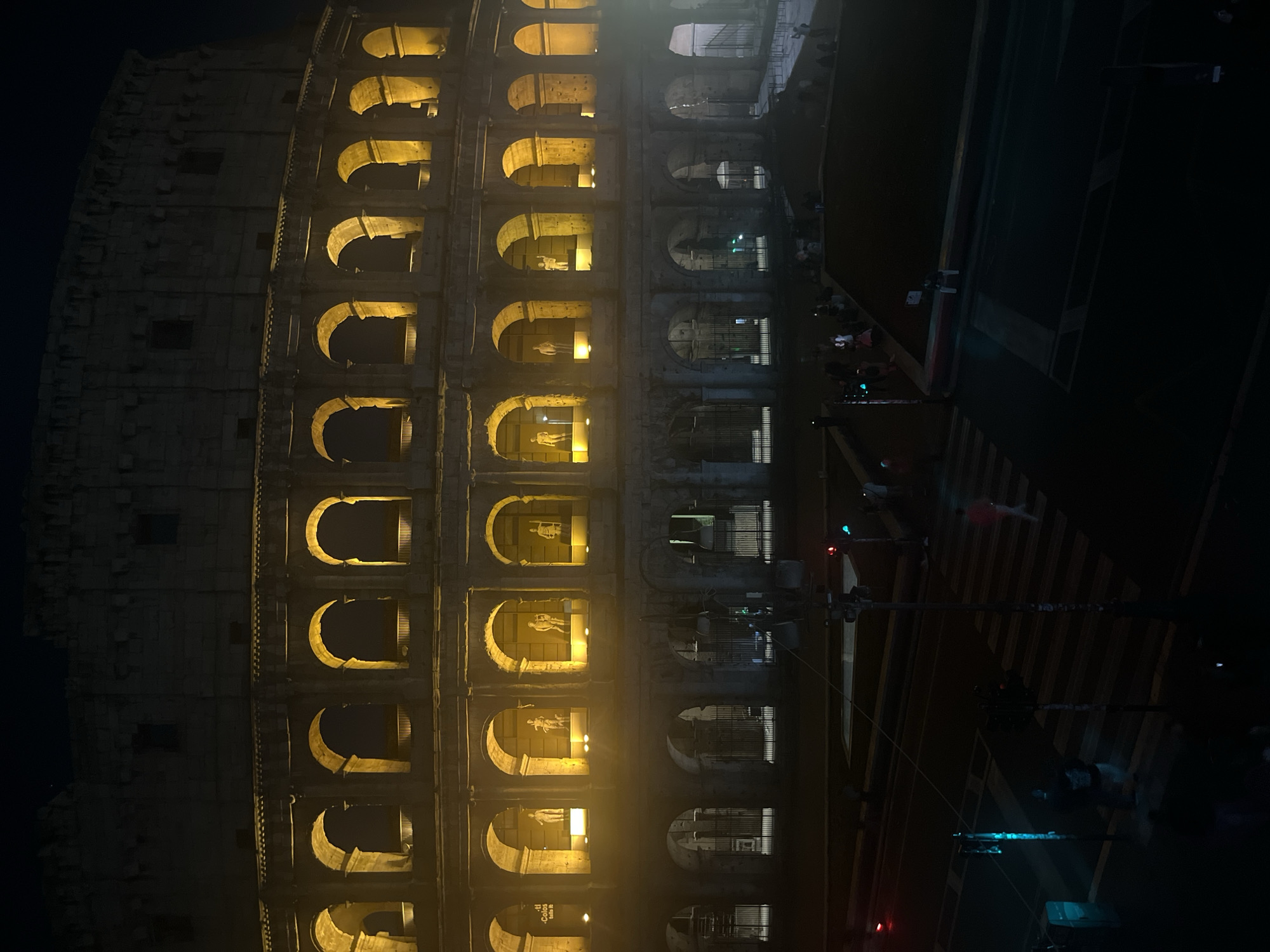 This vertical photograph captures the ancient Colosseum in Rome, Italy, at night. The massive stone amphitheater dominates the left and center, its iconic facade illuminated by warm, yellow-orange lights emanating from within its numerous arched openings. The deep black sky provides a stark backdrop, making the illuminated arches and their textured stone framework stand out vividly. Within several of the brightly lit lower arches, white statues or figures are distinctly visible, displayed for viewing. Some arches to the right show protective metal grates.
To the right of the Colosseum, a modern street scene unfolds with a dark road, featuring a prominent white-striped crosswalk. Traffic light poles equipped with red and green signals are visible, indicating an active urban environment. Several blurry figures of pedestrians can be seen on the sidewalk and near the crossing, suggesting ongoing public activity even after dark. The dramatic artificial lighting emphasizes the Colosseum's grandeur, creating a sense of awe and timelessness, even amidst the contemporary street infrastructure. A partial text "- Colos" is visible inside one arch, referring to the Colosseum. The scene blends historical magnificence with the energy of a city at night.