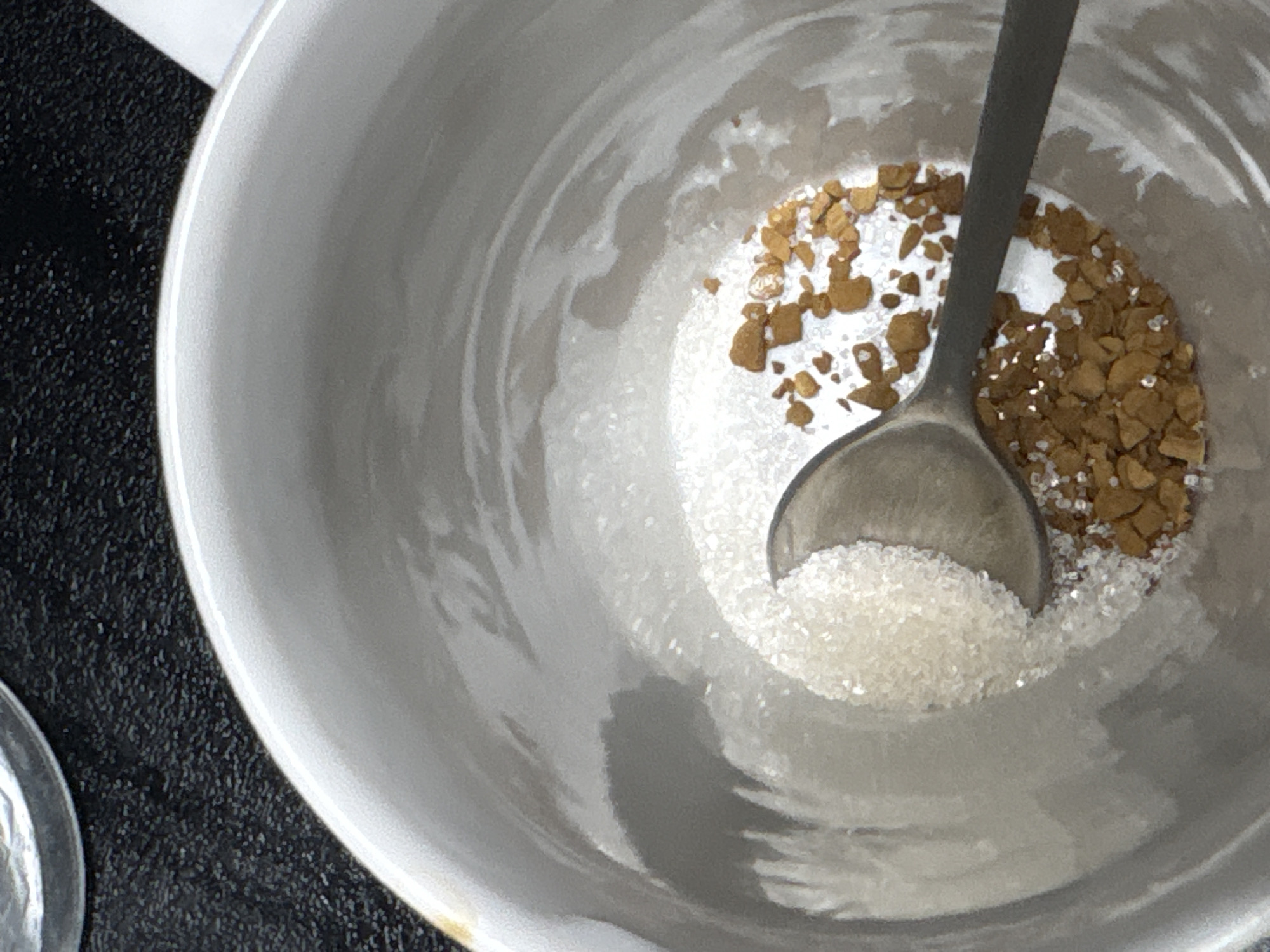 This is a close-up, high-angle shot of instant coffee granules and white sugar in a white ceramic mug. A metal spoon is resting in the mug, scooping up a portion of the sugar. The ingredients are in separate piles, with the sugar on the left and the coffee granules on the right, partially overlapping. The mug is placed on a dark, textured surface, possibly a table. The lighting suggests an indoor setting. There is no visible text in the image. The focus is on the ingredients and the spoon within the mug, giving a sense of preparation for making a hot beverage. The image does not provide any explicit cues about the time of day, weather, or specific emotions. The location context of Mikhaylovsk, Russia, is external information not derivable from the visual content itself.