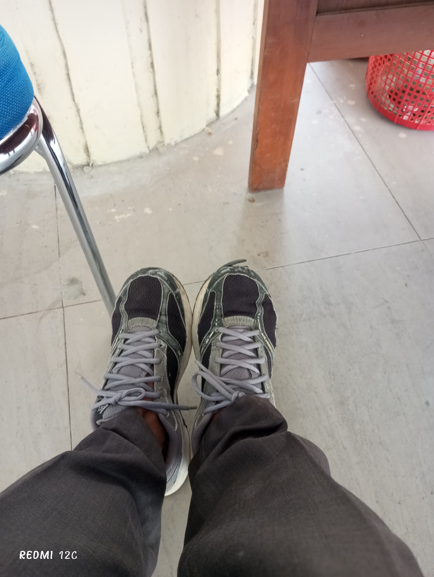 A first-person perspective shot from above shows a person wearing gray trousers and two sneakers. The sneakers are gray with black detailing. The laces are gray and tied. The background consists of a tiled floor, a white wall, the leg of a wooden chair, and part of a blue chair. A red plastic basket is partially visible near the chair. The image includes "REDMI 12C" in the lower left corner. There is no explicit indication of Lhokseumawe, Indonesia.