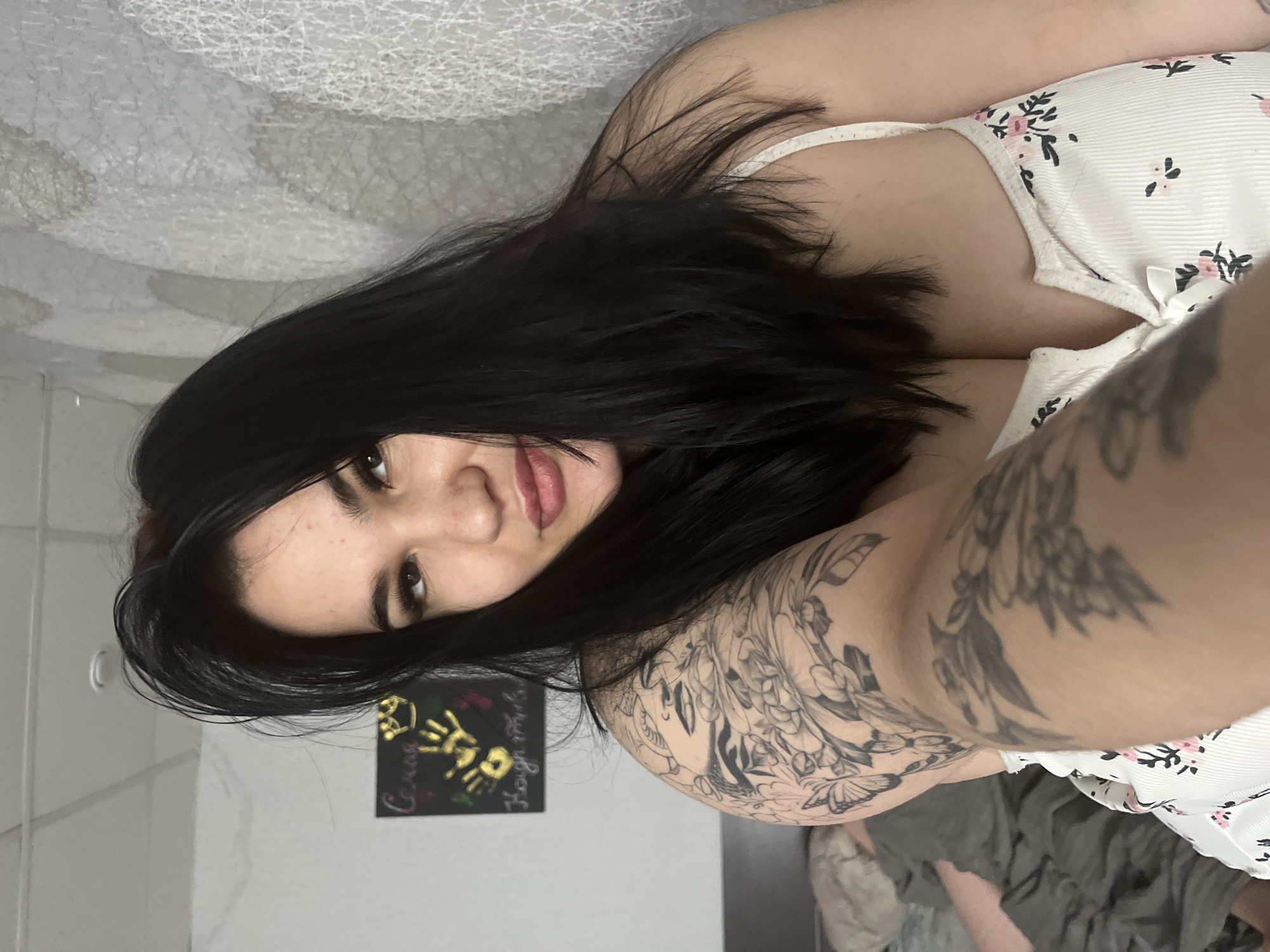 The image features a young woman, likely in her late teens or early twenties, lying down indoors, possibly on a bed or sofa. She is positioned on her side, looking directly at the camera with a direct gaze and a subtle, neutral expression. Her long, straight dark hair frames her face and extends over her shoulders and chest. She has visible dark eyelashes and full lips.
She is dressed in a light-colored, possibly white, strappy top adorned with a repeating pattern of small pink and black flowers. A prominent and detailed tattoo covers her right arm and shoulder, showcasing intricate floral designs and what appear to be butterfly motifs, rendered in black and grey tones.
The background indicates an indoor setting, with a light-colored wall, possibly white or light grey, featuring a subtle textured pattern. A dark rectangular piece of wall art is visible behind her head, on which light-colored text is legible, including the words "Crazy" and "Proud," along with several golden handprints and some colorful scribbles or designs. The bedding or surface she is resting on is a muted grey color. The lighting is ambient indoor light. The setting provides no specific visual cues to identify the location as Chaikovsky, Russia, other than the given context. The primary activity is posing for the camera.