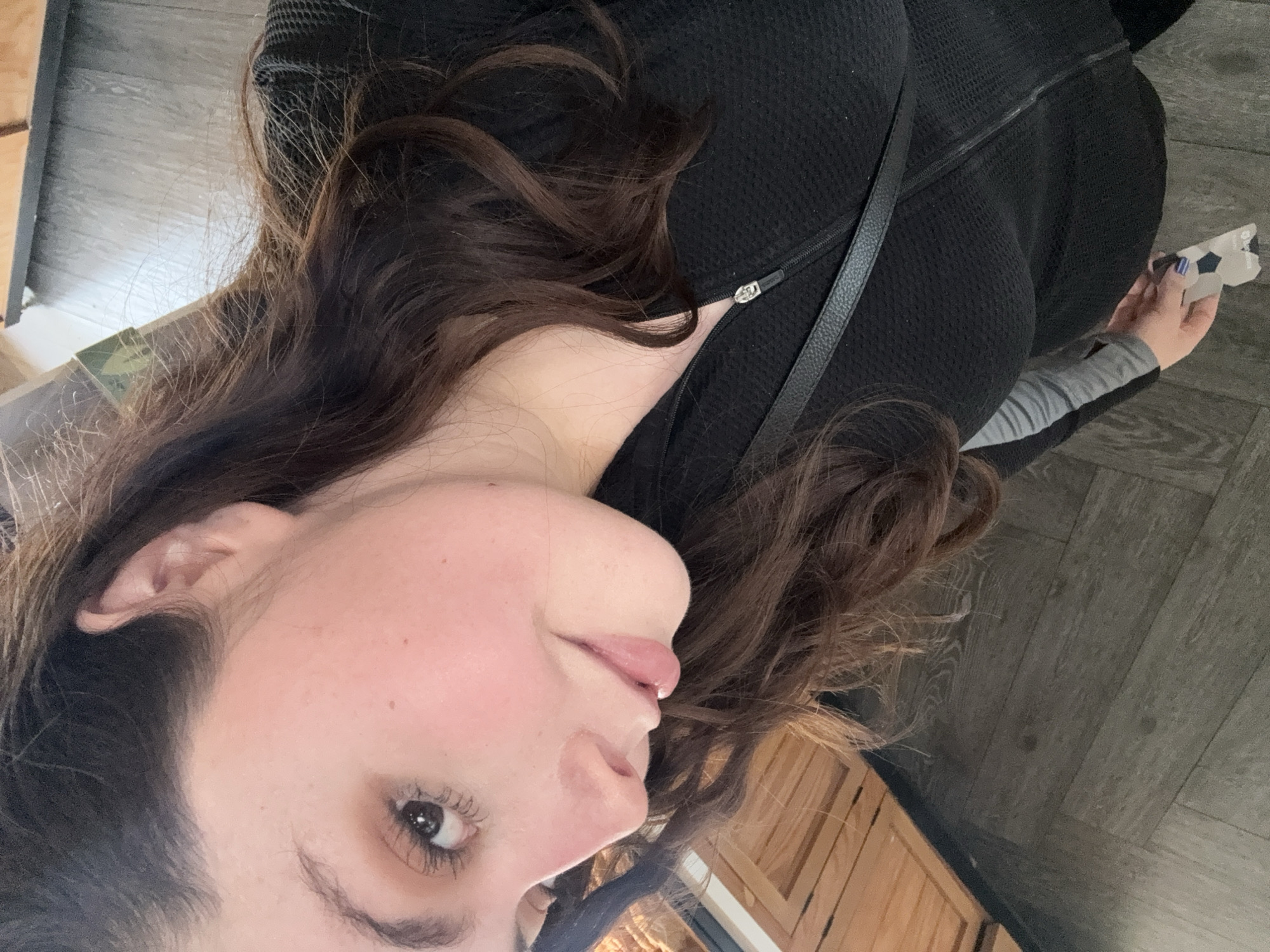 A medium shot depicts a woman with long, dark brown hair. She is wearing a black quilted jacket with a thin black strap across her chest. Her left hand is visible, holding a small, white object with black geometric designs. She appears to be indoors, with a partial view of light-colored wooden cabinets and gray flooring in the background. The location is not discernible from this image.