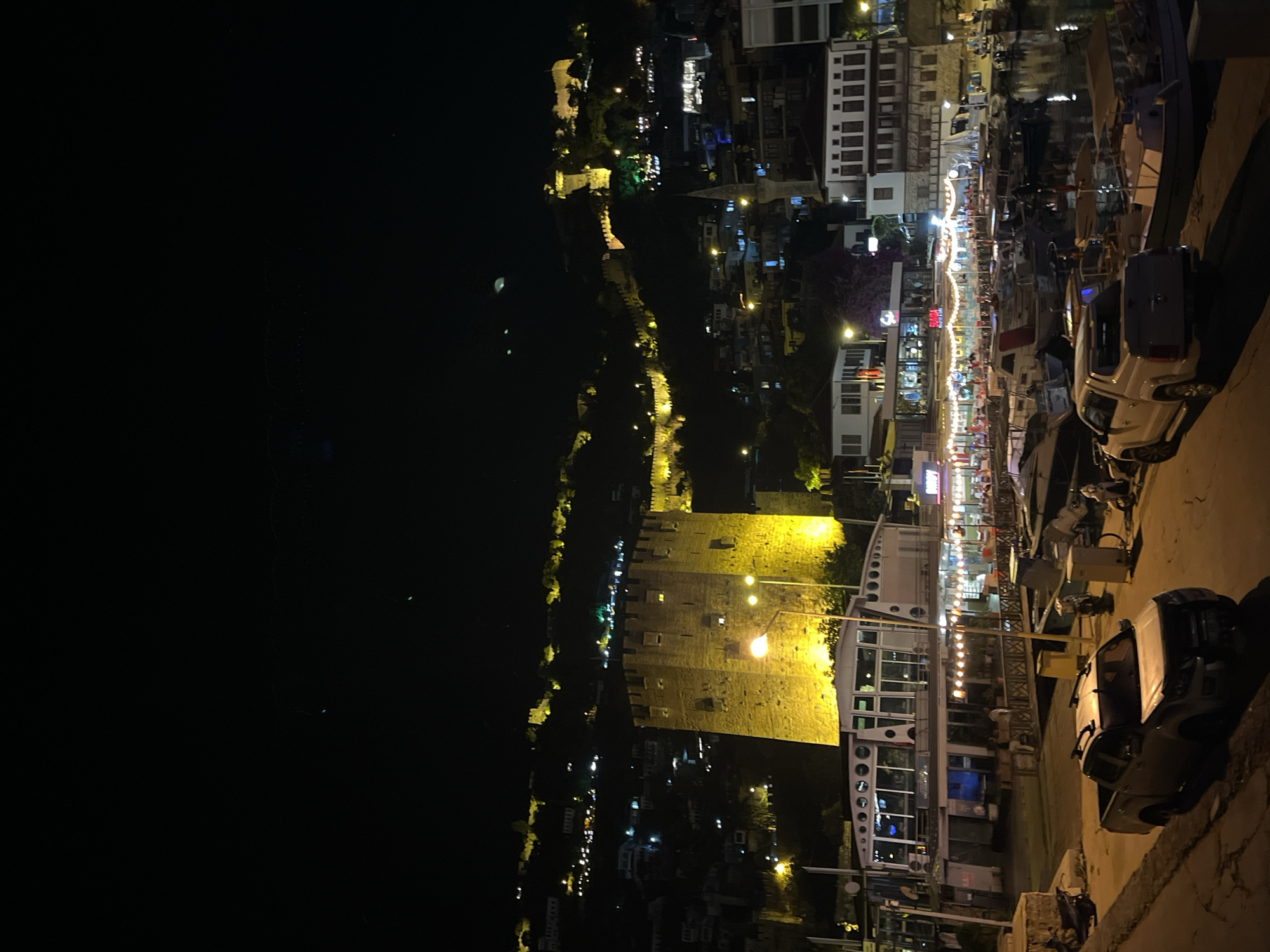 This is a nighttime photograph of a vibrant coastal town in Alanya, Turkey, showcasing a bustling harbor area and an illuminated historical structure. The image is filled with artificial lights against a dark sky and water, creating a lively atmosphere.
On the left side of the frame, a dark expanse of water, likely the sea or a large harbor, stretches out with subtle reflections of distant lights. Along the water's edge, a long, ancient-looking fortified wall, possibly part of a castle or city defenses, is brightly lit with warm yellow light. A prominent, large square or rectangular stone tower, also intensely illuminated in yellow, stands near the water, dominating the mid-left section of the image.
To the right, a lively waterfront promenade is visible, bustling with activity. Numerous buildings, likely restaurants and shops, line the street, adorned with string lights and various glowing signs. Boats are docked along the quay, and their outlines are visible amidst the reflections on the water's surface closer to the promenade. In the immediate foreground on the right, a paved road or path is visible, with two vehicles parked: a white pickup truck and another darker vehicle behind it.
The scene suggests a popular evening destination, with people likely enjoying dining, walking, and the ambiance. One visible text cue, "RUMBA," appears on a sign for what seems to be an establishment along the promenade. The overall impression is one of a picturesque and active port city illuminated at night.