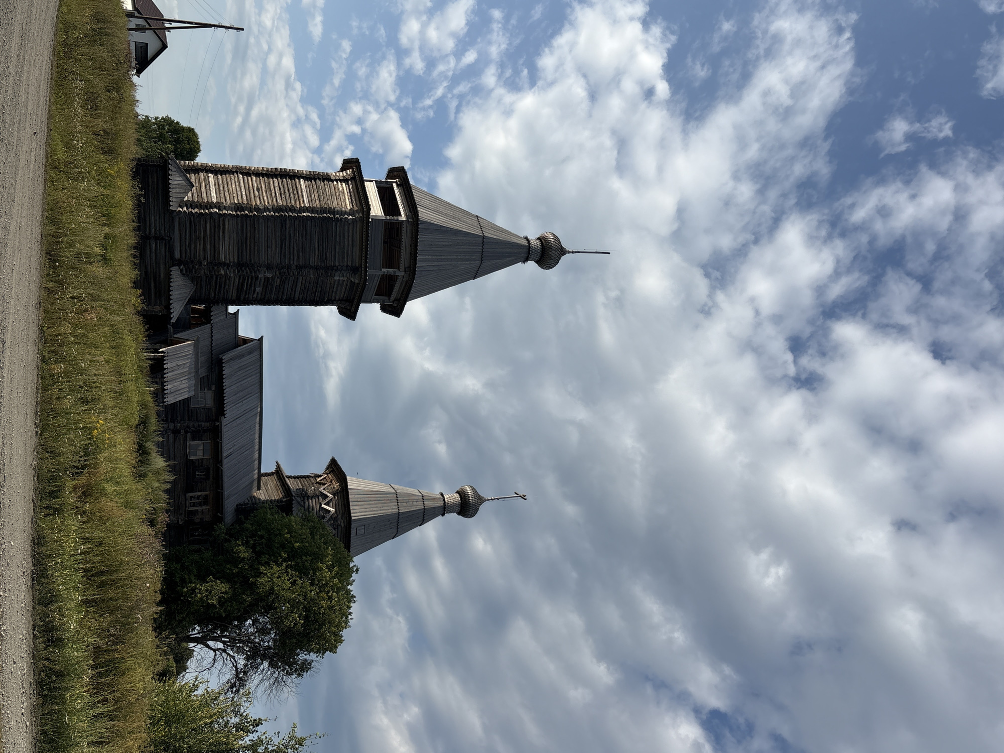 This is a vertical, medium-shot, outdoor photograph of two wooden churches in the village of Tipinitsy, Russia, under a partly cloudy sky. The church on the left has a tall, square, wooden bell tower with an octagonal, conical roof topped with a finial. The church on the right has a smaller, octagonal tower, also with a conical roof topped with a cross. Both structures are made of dark, weathered wood. In the foreground, there is a dirt road and tall, dry grass. Behind the churches, there is a dense green tree and other vegetation. A power pole and a small white building are visible in the distance to the left. The lighting suggests it is daytime, possibly late afternoon, with the sun casting some shadows.
