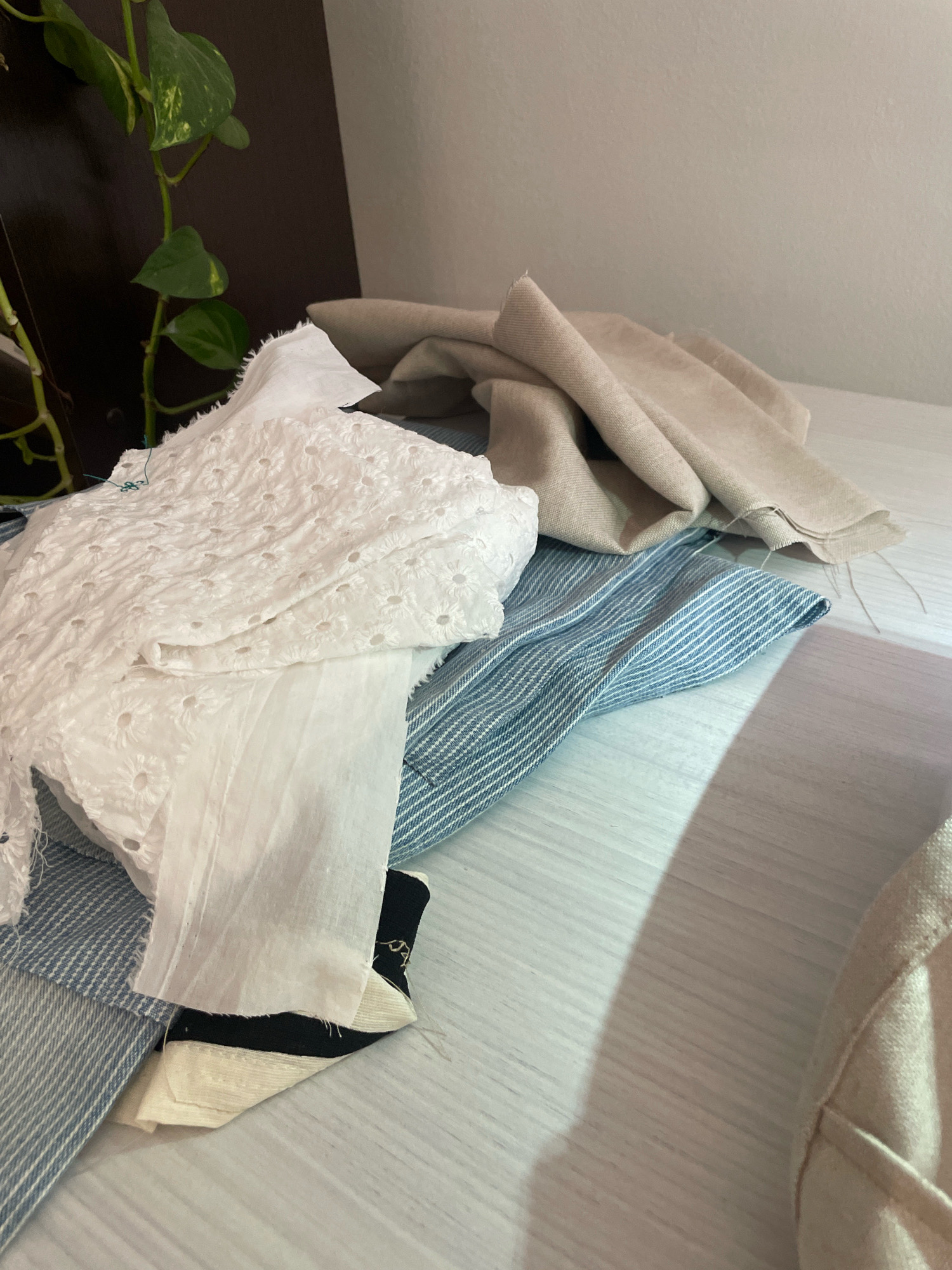 The image shows a pile of fabric scraps on a light-colored, textured surface that resembles wood. The fabrics include white eyelet lace, a blue and white pinstriped fabric, and a beige linen-like material. There are also smaller pieces of black and cream colored fabric visible at the bottom. A dark-colored plant with green leaves is visible in the upper left corner, partially obscuring a dark vertical surface. The lighting suggests an indoor environment with a shadow cast on the surface from the upper right. There is no indication of the city of Atarfe, Spain, or any specific location within it.