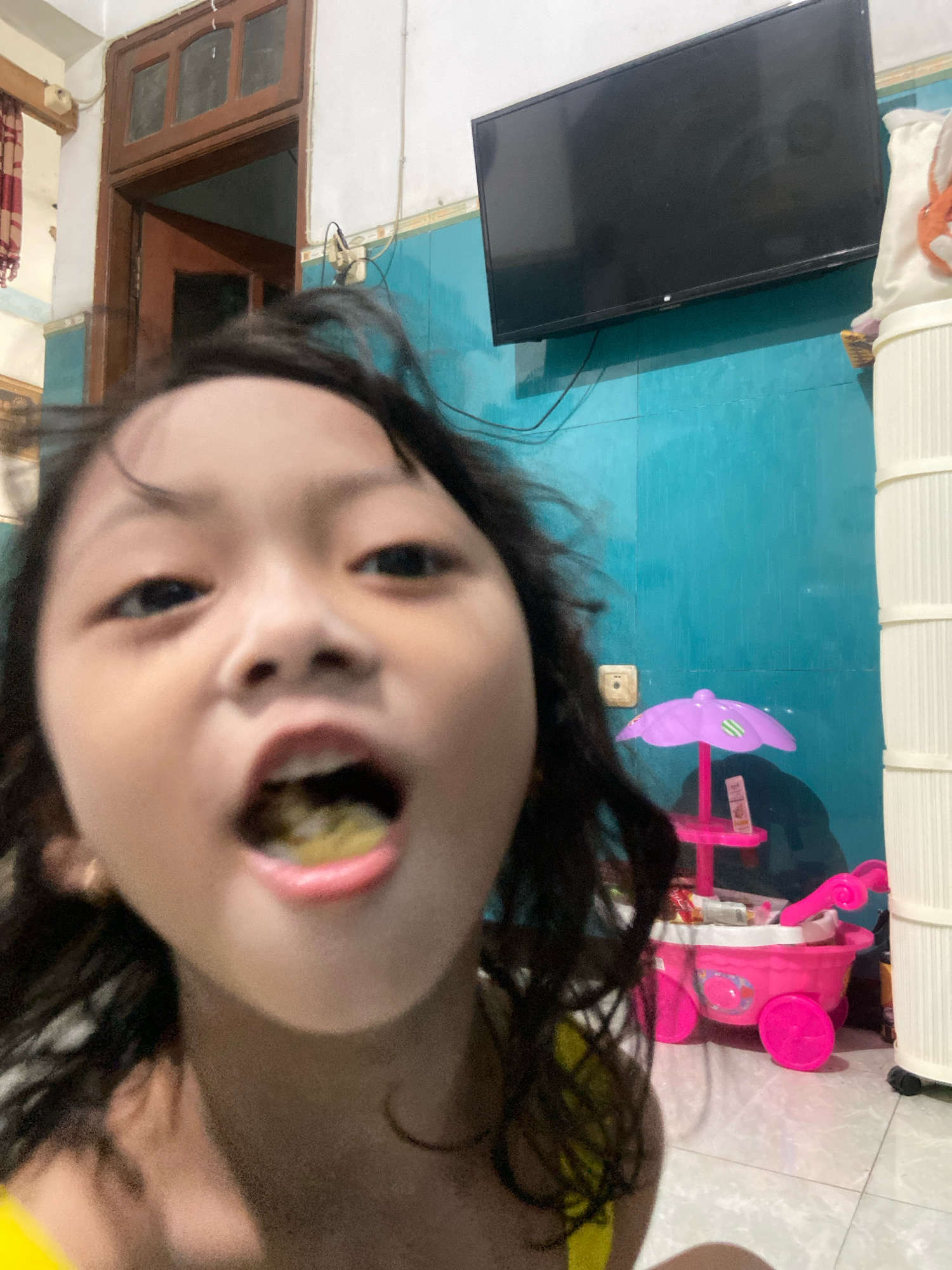 The image shows a close-up of a young girl with dark hair, her mouth open as if eating or speaking. She is indoors, with a teal-colored wall behind her. To the left, a wooden door with glass panes is visible. A black flat-screen television is mounted on the wall above the door. To the right, a white ribbed cylindrical object, possibly a laundry hamper, stands next to a pink toy stroller with a purple umbrella. The floor appears to be tiled. The image is not specific to Gelam, Indonesia, but rather depicts a common indoor domestic setting.