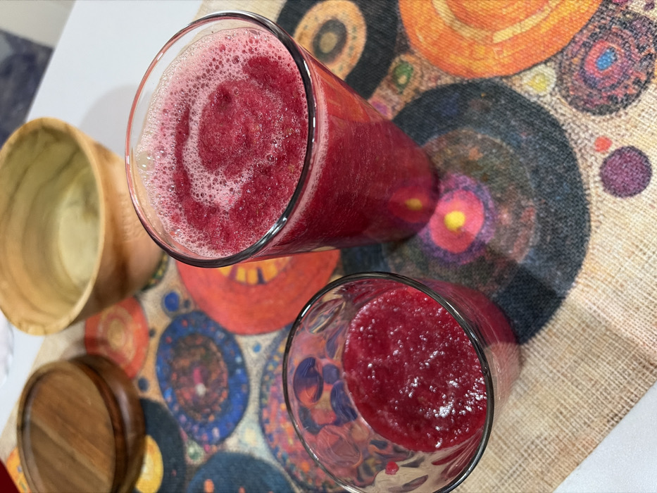 The scene, located in Igboefon, Nigeria, displays two clear glass vessels containing a dark red, frothy beverage, likely a smoothie or juice, viewed from an overhead perspective. A tall glass, positioned right of center, is nearly full, exhibiting a distinct frothy swirl on its surface. To its lower left, a shorter glass contains approximately one-third of the same liquid, which appears slightly granular. These glasses are situated on a woven fabric table runner adorned with multiple concentric circular patterns in vibrant hues of red, blue, orange, purple, and yellow. In the upper background, partially blurred, are a light-colored wooden bowl and a darker wooden lid, resting on a white surface adjacent to the patterned runner. No individuals or active interactions are depicted within the frame.