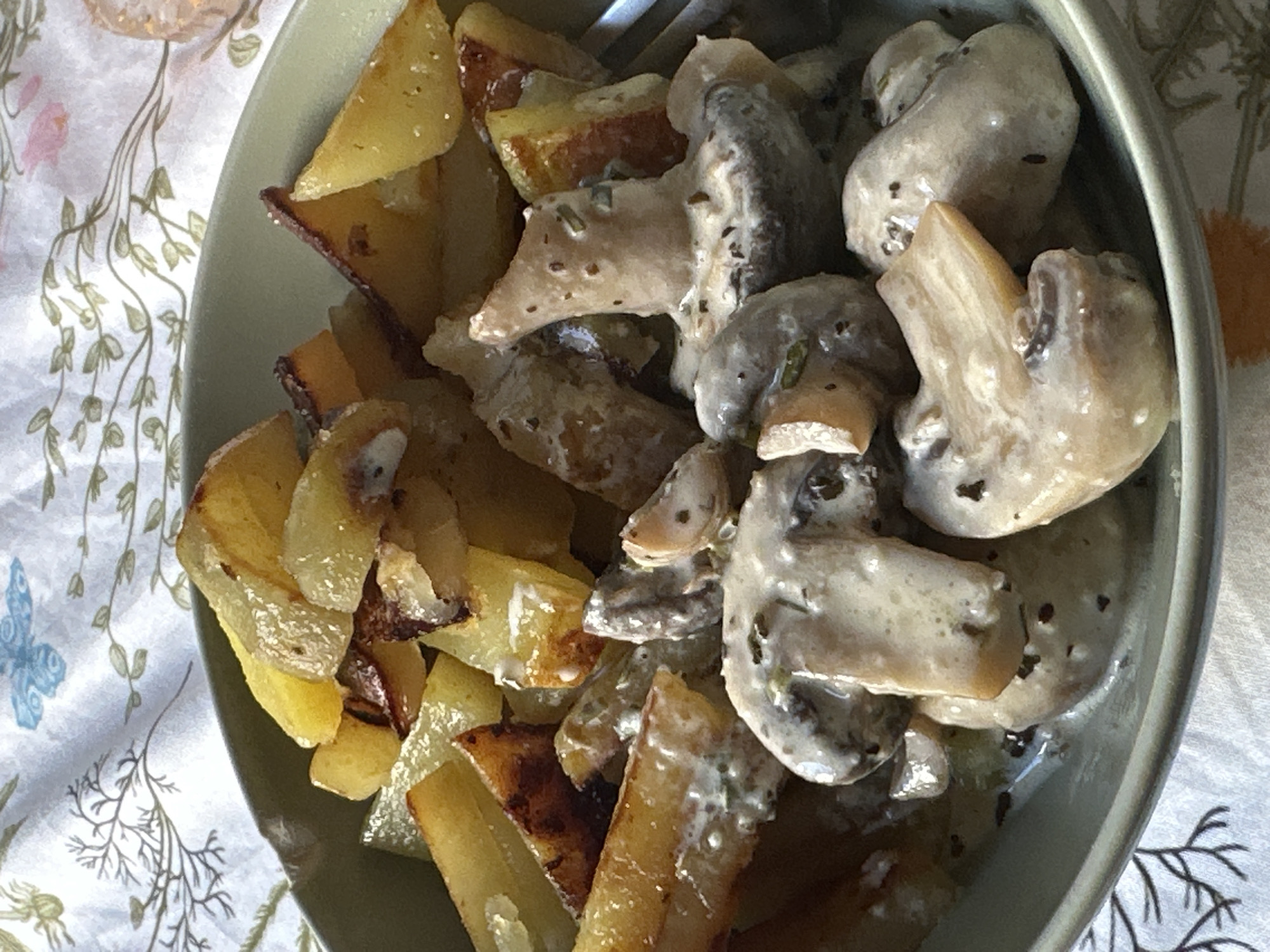 A close-up shot presents a bowl filled with a meal consisting of fried potatoes and mushrooms in a creamy sauce. The potatoes are golden brown, suggesting they have been fried to a crispy texture. The mushrooms are coated in a light-colored sauce, speckled with herbs. The food is contained within a gray-green bowl. The scene is set on a patterned tablecloth, featuring floral and butterfly motifs. The image does not explicitly specify the location in the city of City of Zagreb, Croatia, and there are no people visible. The focus is primarily on the food and its presentation.