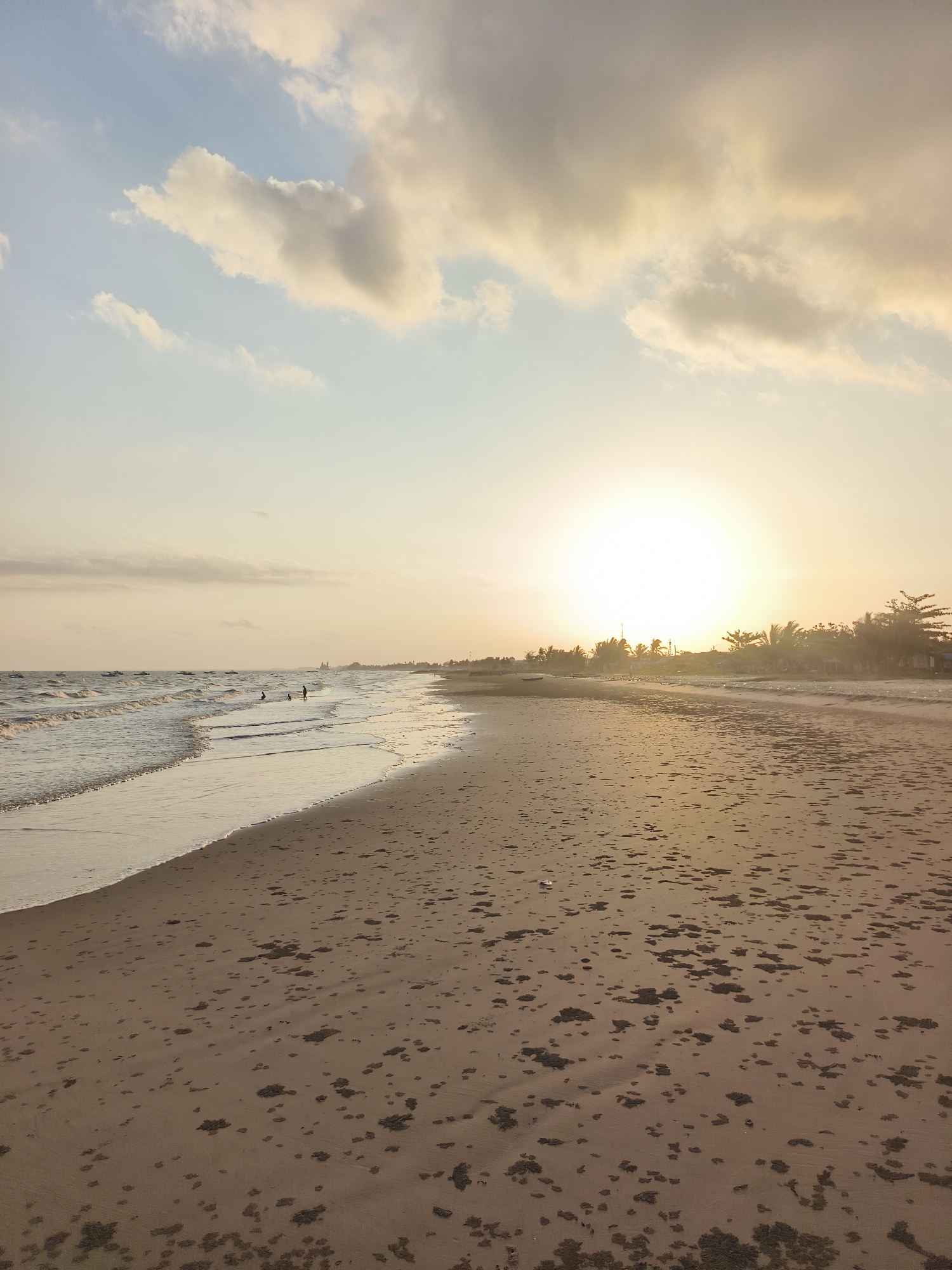 The image depicts a beach scene at Kupang Berkah Jaya, Indonesia, during what appears to be sunset. The sun, a bright, glowing orb, is positioned slightly to the right of the frame, casting a warm, golden light across the sky and the beach. Wispy clouds, tinged with orange and yellow hues, are scattered across the pale blue sky.
The foreground is dominated by the sandy beach, which is textured with dark, scattered patches that could be seaweed, debris, or damp sand. The wet sand at the edge of the water reflects the sky's colors. Gentle waves are rolling in from the left side of the image, creating a foamy, white edge where they meet the sand.
In the distance, along the shoreline, a line of trees and vegetation is visible, silhouetted against the bright sky. There are also some structures that appear to be buildings or huts. Several small boats are dotted on the water further out.
A few small figures, likely people, are present on the beach and in the shallow water. One person is standing near the water's edge, and another is wading. Further down the beach, other figures are also visible. The overall atmosphere is tranquil and serene, typical of a beach at the end of the day.