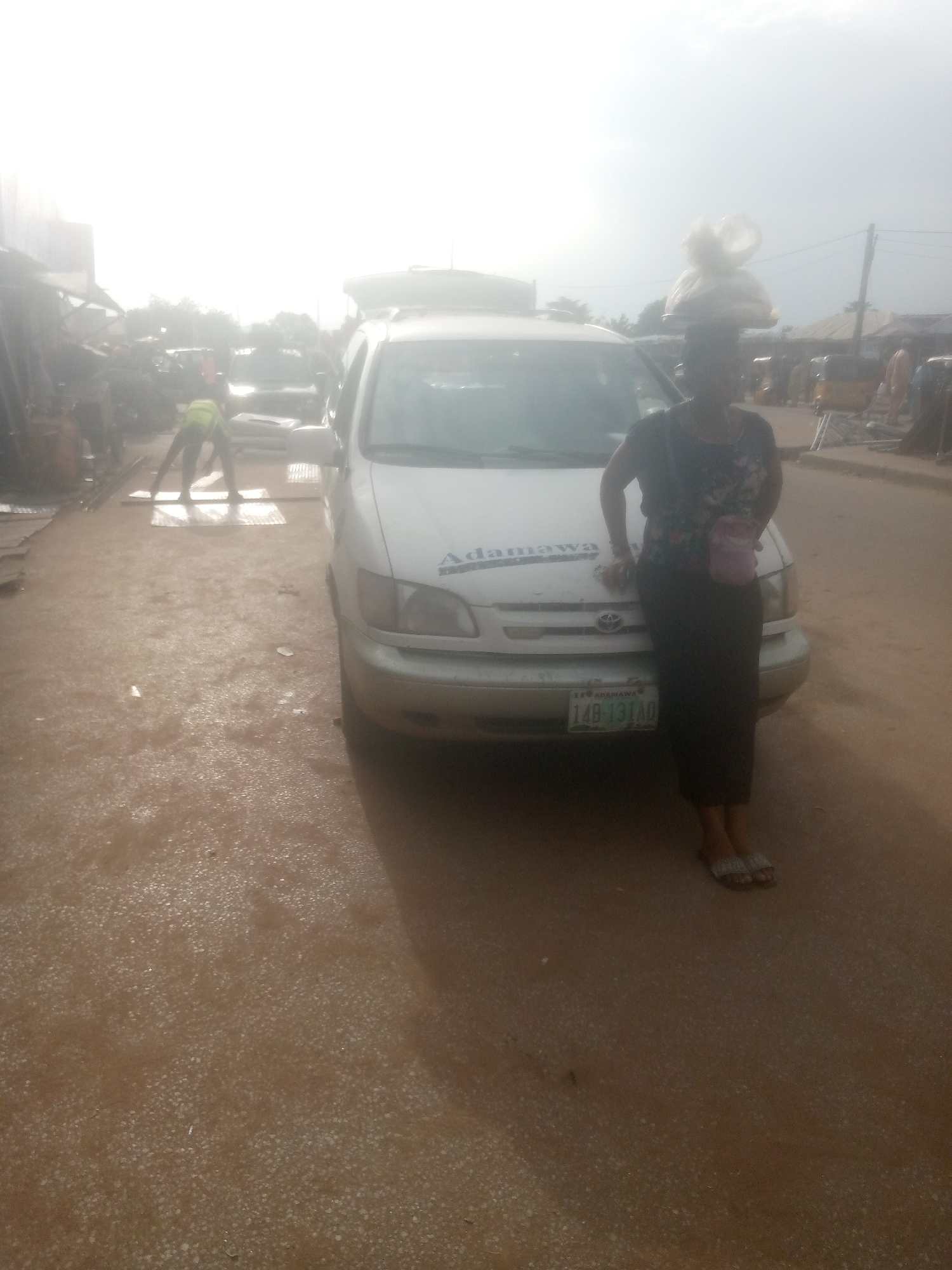A woman stands next to a white van, with a large bundle of items balanced on her head. She wears a dark, floral-patterned shirt and a long, dark skirt. A small pink purse is slung across her body. Her feet are adorned with silver, jeweled sandals. The van has the word "Adamawa" written on its hood and a license plate that reads "148-131AD". In the background, another person in a bright yellow vest is bent over, seemingly handling some corrugated metal sheets on the ground. The scene appears to be a street or market area, with other vehicles and structures visible in the distance. The sky is bright, suggesting it is daytime, possibly late morning or afternoon. The ground is dirt and appears wet or muddy in places, with reflections visible, indicating recent rain or damp conditions. The overall atmosphere seems to be that of a busy, everyday scene in a Nigerian town.