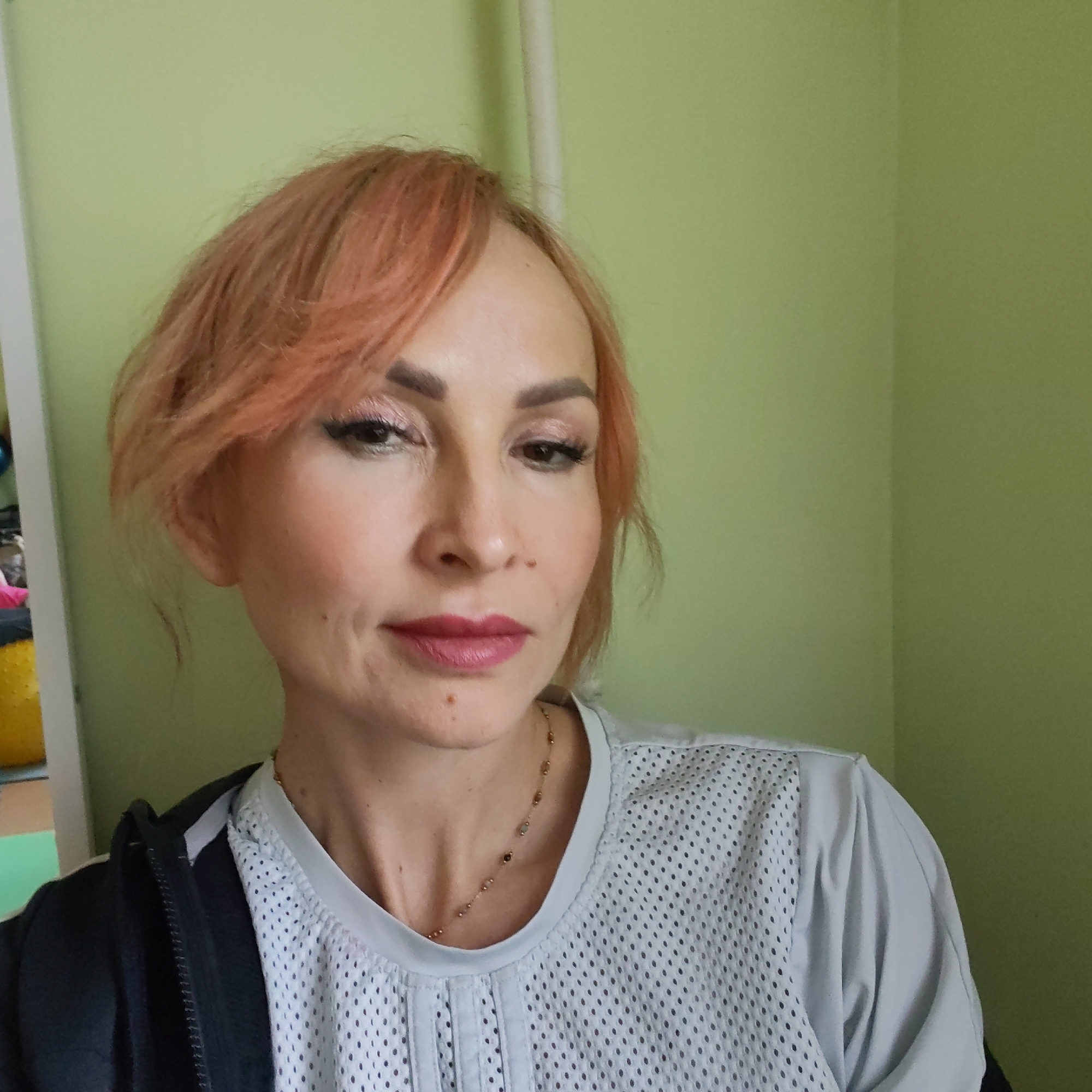 A close-up portrait captures a woman with reddish-blonde, chin-length hair, styled with bangs sweeping to her right. She appears to be in her 30s or 40s, with a fair complexion. Her makeup includes subtly defined eyebrows, eye shadow, eyeliner with apparent false lashes, and a rich, dark pink lipstick. A small mole is visible above her upper right lip and another on her chin.
She is wearing a light grey or off-white t-shirt made of a mesh-like, perforated fabric, layered under a dark (likely black) zip-up jacket. A delicate, gold-toned necklace with small, scattered beads is visible around her neck. The woman gazes slightly downwards and to her right, conveying a calm and somewhat pensive expression.
The background consists of a plain, light green wall. To the far left, a vertical white frame or pillar is partially visible, alongside blurred hints of other objects, possibly including a yellow exercise ball, suggesting an indoor setting. The lighting is soft and even, typical of an indoor daytime photograph. The scene is set in Chaikovsky, Russia. There is no discernible text within the image.