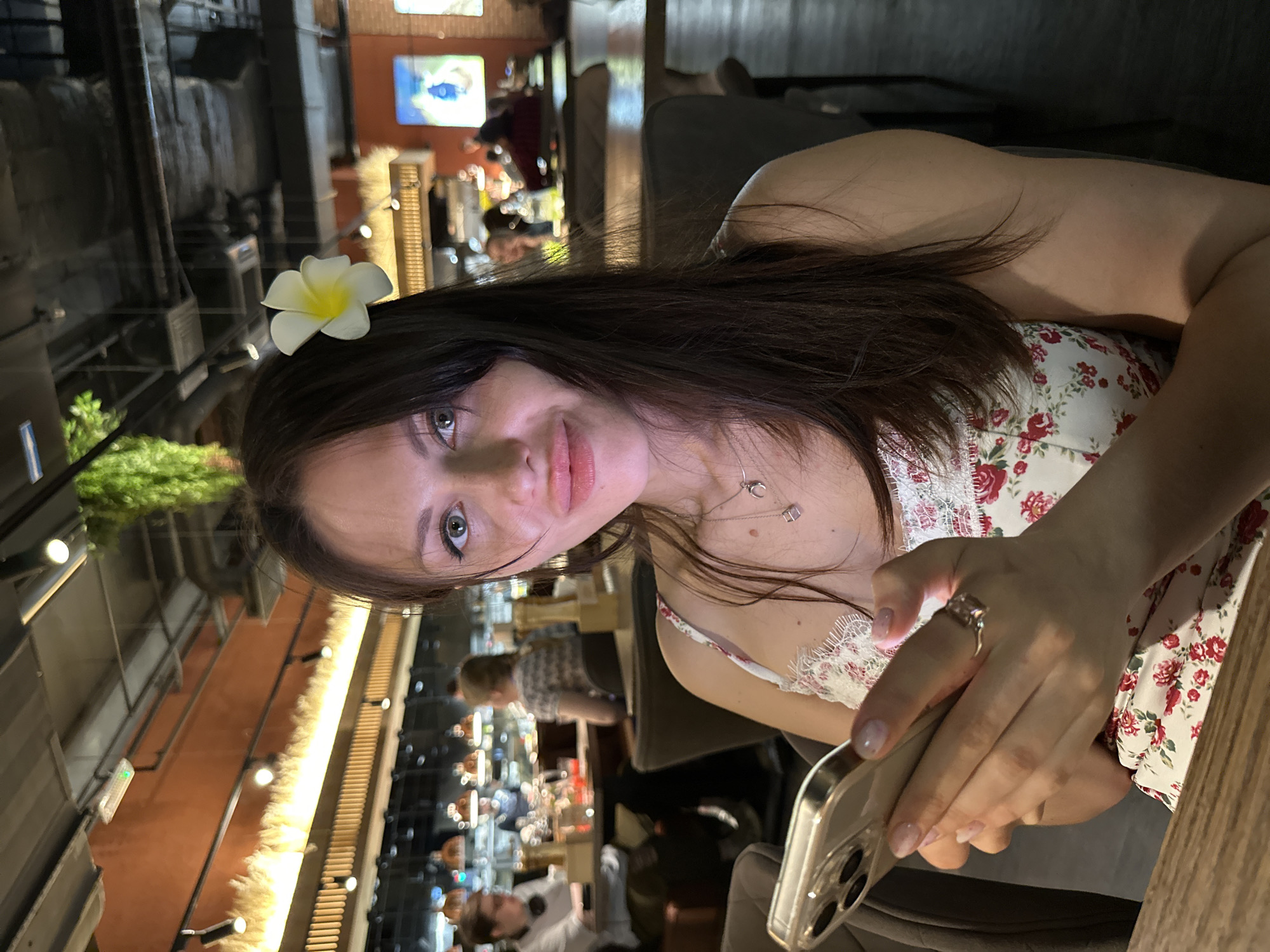 A woman with long brown hair and a white flower behind her ear is seated in what appears to be a restaurant. She is holding a smartphone in her hands. The restaurant interior features a partially visible bar area with bottles and shelves, suggesting it is located indoors. Other people are seated at tables in the background, indicating an occupied dining area. The overall image suggests an evening or night setting, potentially a casual or social occasion at a restaurant. It is not possible to determine from this picture the location in Saint Petersburg, Russia.