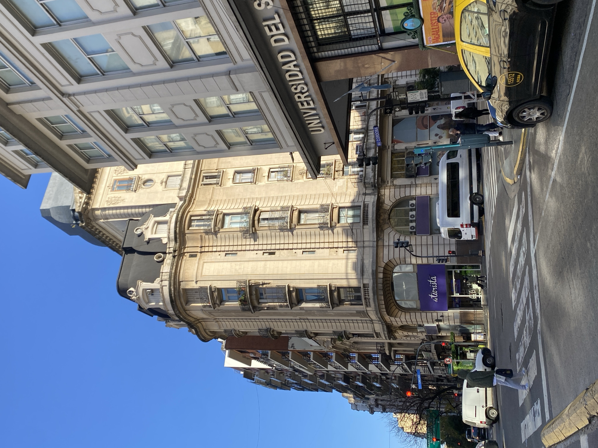 The image shows a street scene in Buenos Aires, Argentina, with a focus on the architecture. The Universidad Del Salvador building dominates the left side, with its detailed facade and signage. The street is busy, with pedestrians crossing at a zebra crossing. A yellow and black taxi is parked on the right side of the street. Several white vans are also present, indicating potential commercial activity or deliveries. Street signs and traffic lights are visible, along with a glimpse of other buildings in the background. The sky is clear and blue.