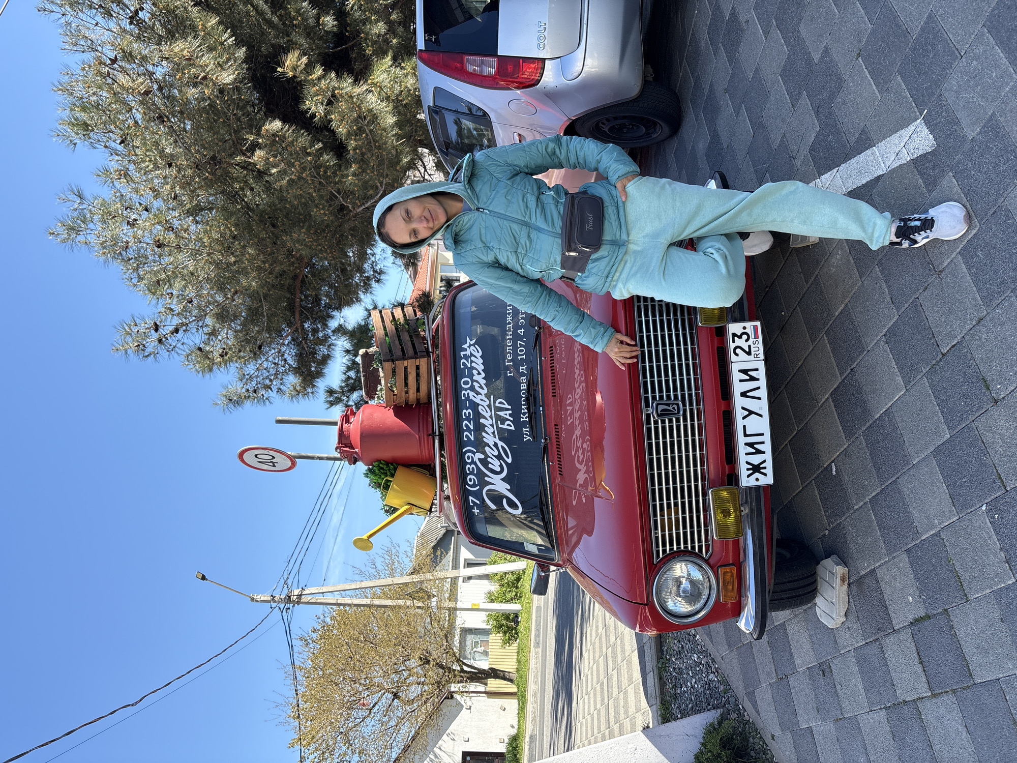 A woman poses leaning on a red vintage car parked on a paved street under a clear blue sky. She is light-skinned, with a slight smile, and is wearing a light blue/mint green hooded tracksuit, white and black athletic shoes, and a black waist bag with "Trust" written on it. Her right hand rests on the car's hood, and her left leg is bent.
The car is a red Zhiguli (Lada), featuring a chrome grille and headlamps. On its roof rack, there are wooden crates, a red jerry can, and a yellow watering can. The windshield has text in Russian, indicating "Жигулевские БАР" (Zhigulevskie BAR), a phone number (+7 (939) 223-30-2xx), and an address: "г. Геленджик ул. Кирова д. 107, 4 эт" (Gelendzhik, Kirova street, house 107, 4th floor). The front license plate displays "ЖИГУЛИ" and "23 RUS", confirming the vehicle's make and region (Krasnodar Krai, Russia).
Behind the red car, a silver Mitsubishi Colt is parked, partially visible, with "COLT" written on its rear. The street features dark grey paver stones. In the background, large evergreen trees with cones and other trees are visible, along with utility poles, power lines, and a "40" speed limit sign. The bright sunlight suggests it is daytime with good weather.