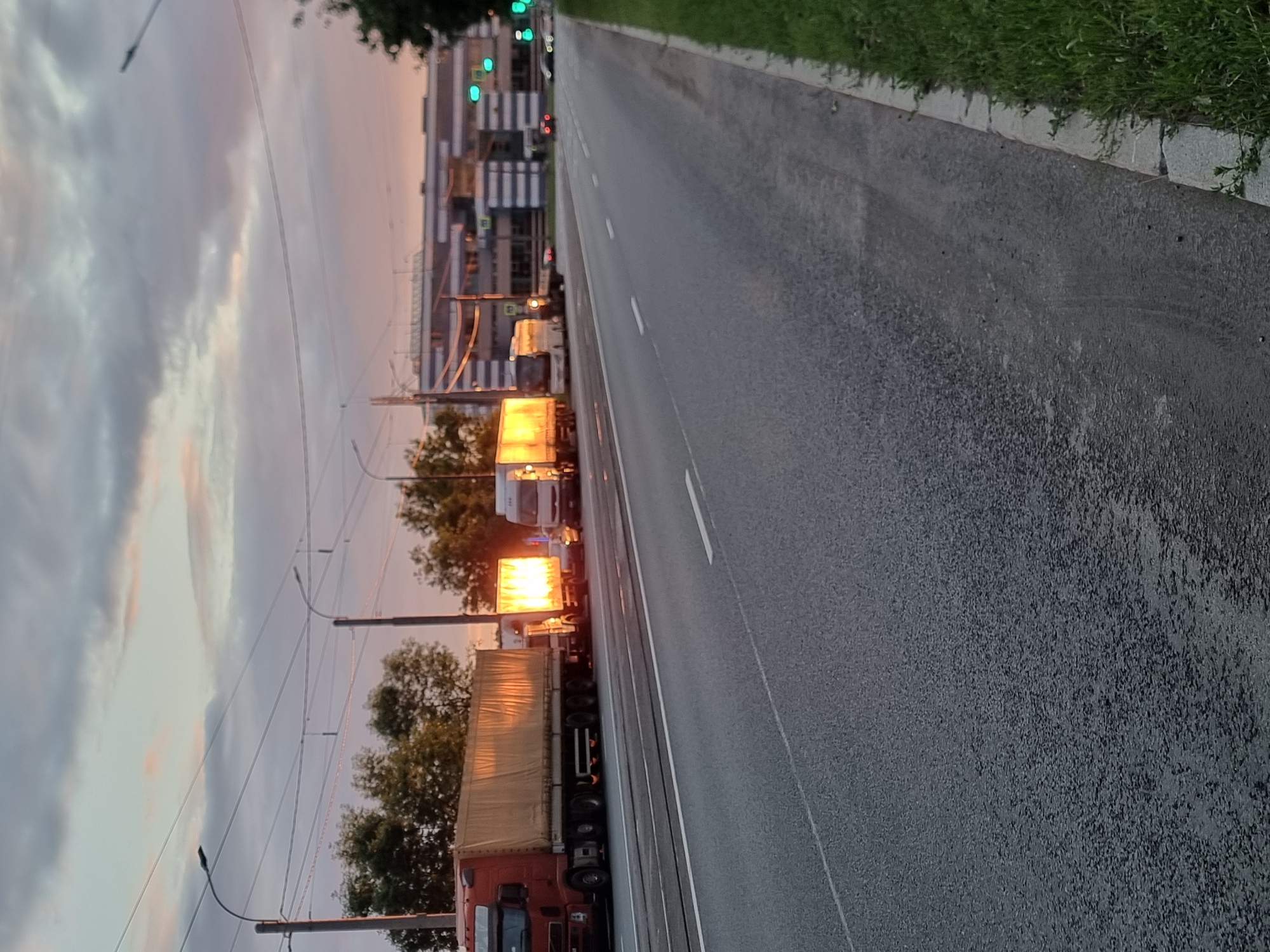 The image depicts a road in Saint Petersburg, Russia, likely during dusk, given the twilight sky with pink and gray clouds and the illuminated headlights and trailer lights of vehicles. Several large trucks, some with canvas-covered trailers, are parked in a line on the right side of a multi-lane asphalt road. One truck has its headlights on, illuminating its trailer. Further down the road, traffic lights are visible, and other vehicles, including cars and possibly smaller trucks, are in motion. A modern, multi-story building with a striped facade is visible in the background. The road is bordered by a grassy verge on the right. The scene suggests a possible traffic congestion or a temporary stop for these vehicles.