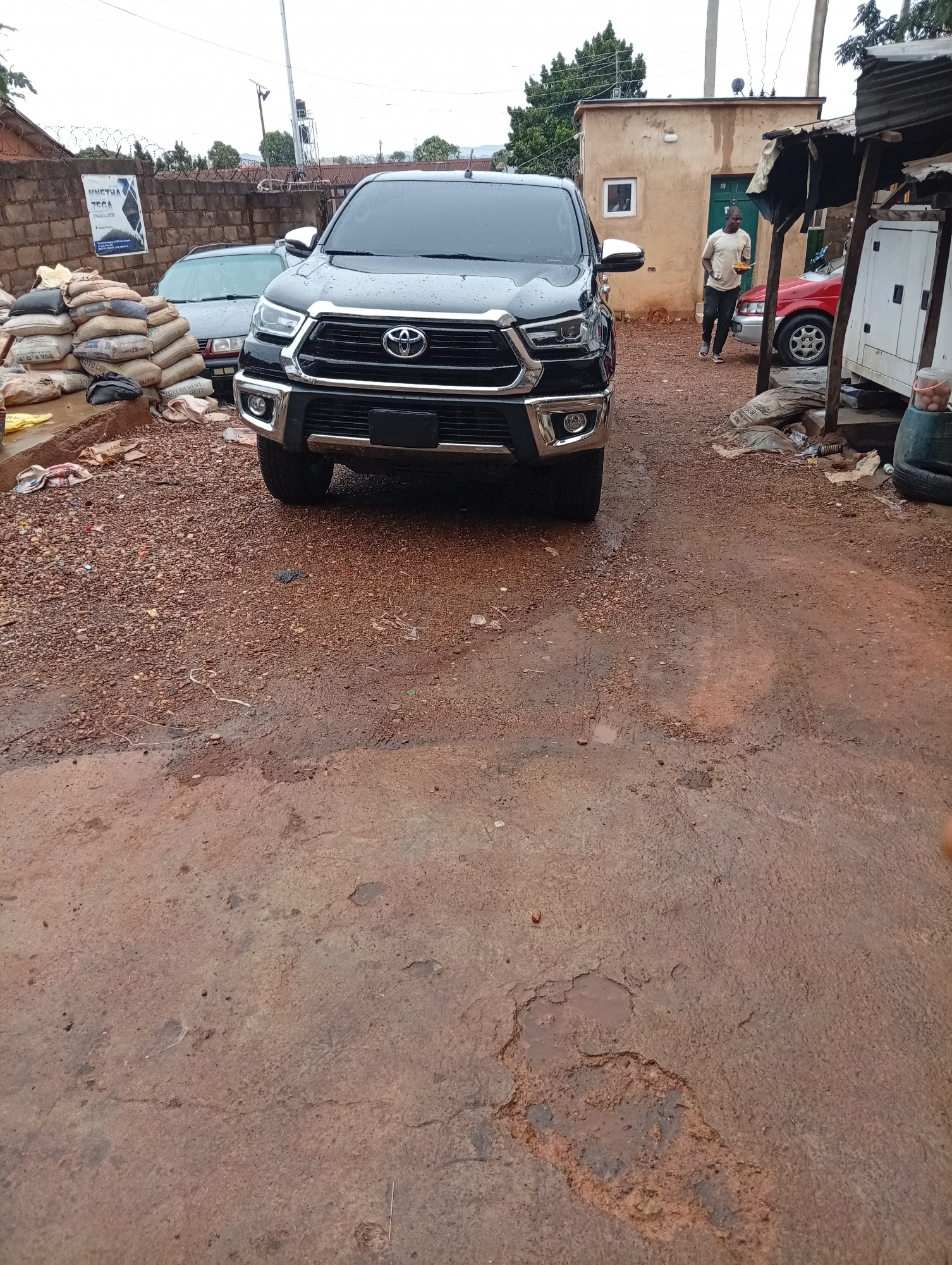 The image depicts a black Toyota Hilux pickup truck parked on a dirt and gravel road in Gura Top, Nigeria. The foreground shows a reddish-brown surface with patches and small puddles of water. In the background, there is a wall with barbed wire, bags of cement stacked against it, and other cars parked behind the truck. A man is walking in the background towards a building with a green door, while a generator is to the right of the man. The lighting is overcast, suggesting a wet or cloudy day.