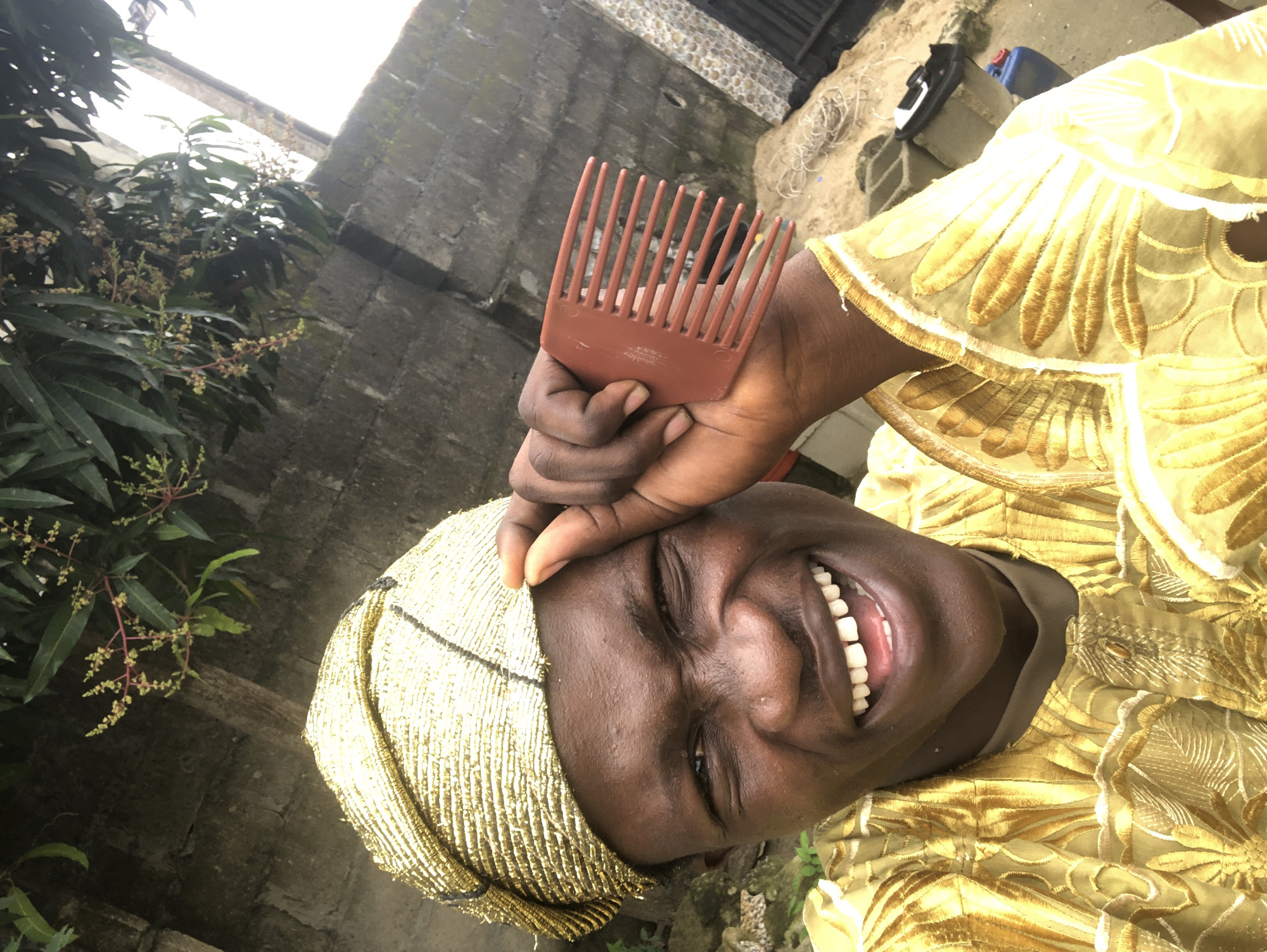 A young Nigerian man in Ado, Nigeria, is taking a selfie outdoors. He is wearing a traditional golden headwrap and a matching golden embroidered robe. He is holding a brown comb to his forehead, and his mouth is open in a wide, joyful smile, showing his teeth. The background shows a brick wall, some greenery from a tree, and a slightly overcast sky.