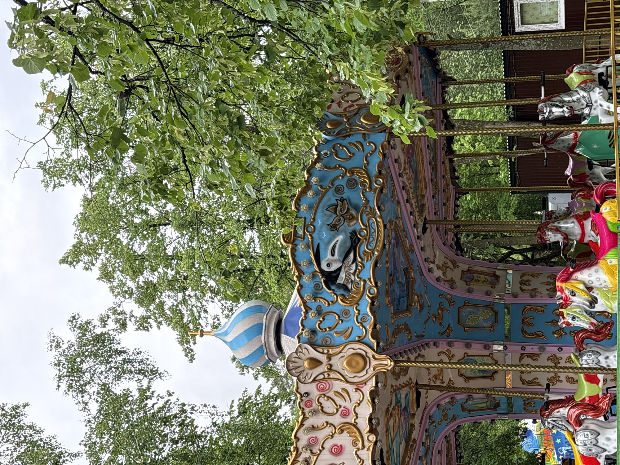 A stationary carousel stands partially obscured by dense green foliage under an overcast sky. The carousel features an ornate, light blue and white striped conical dome topped with a golden finial. Below the dome, the carousel's upper perimeter is decorated with sculpted panels in hues of light blue and pale pink, accented with intricate gold filigree. These panels depict various motifs, including a white swan-like bird with black details, a butterfly, and swirling patterns. The carousel's interior walls are paneled in light blue and pink, showcasing smaller framed illustrations such as a sailboat and another butterfly. Numerous carousel horses are visible, predominantly white and gray, with some featuring red manes and tails, while others have yellow accents and bright green saddles. These horses are supported by thin, twisted golden poles. In the background, dark brown corrugated metal sheeting forms a barrier or building, with a single white-framed window visible. Another colorful amusement ride is partially visible on the far left. The surrounding environment consists of lush, green deciduous trees with varied leaf tones, some appearing lighter green, suggesting recent growth or wetness. This scene is located in Perm, Russia.