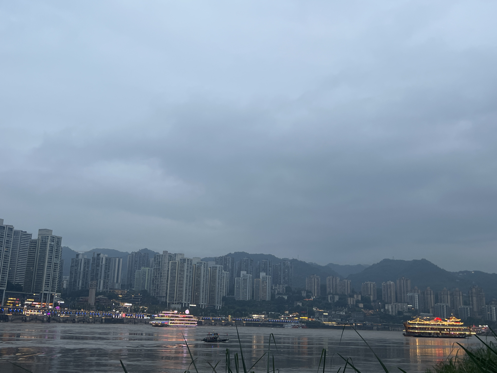 The image depicts a wide river with a city skyline on the opposite bank. The skyline is dominated by numerous tall, modern buildings, many of which are illuminated with lights. In the foreground, the river's surface reflects the lights from the city. Several boats are visible on the water, including a large, multi-story tour boat adorned with numerous lights. In the distance, a range of mountains can be seen behind the city, partially obscured by the overcast sky. The overall atmosphere is somewhat dim, suggesting either early morning or evening, with heavy clouds contributing to the muted light. The location is identified as the Nan'an District in China, implying this is likely a view of the Yangtze River or a major tributary within the Chongqing municipality, given the characteristic mountainous backdrop and dense urban development. No individuals are clearly identifiable within the scene. The image captures a blend of urban infrastructure and natural landscape under a cloudy sky.