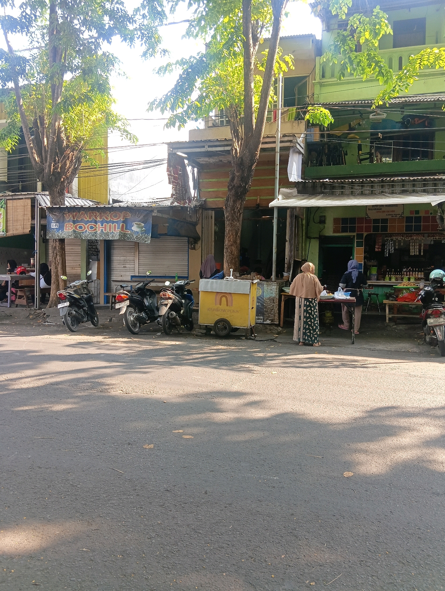 The image depicts a street scene in front of a row of shops in Gapurosukolilo, Indonesia. A large tree with sparse leaves stands on the left, casting shadows on the asphalt road. In front of the shops, three motorcycles are parked. A banner above one shop reads "WARKOP BOCHILL" with a coffee cup icon. A yellow food cart with the words "RAINBOW" and "BOW PUKIS" is also present. Two women are visible near a table in front of one of the shops, with one woman wearing a light brown hijab and a patterned skirt, and the other wearing a dark hijab and trousers. Behind them, bottles and other items are displayed on shelves within the shop. Sunlight illuminates the scene, highlighting the bright green and yellow facades of the buildings.
