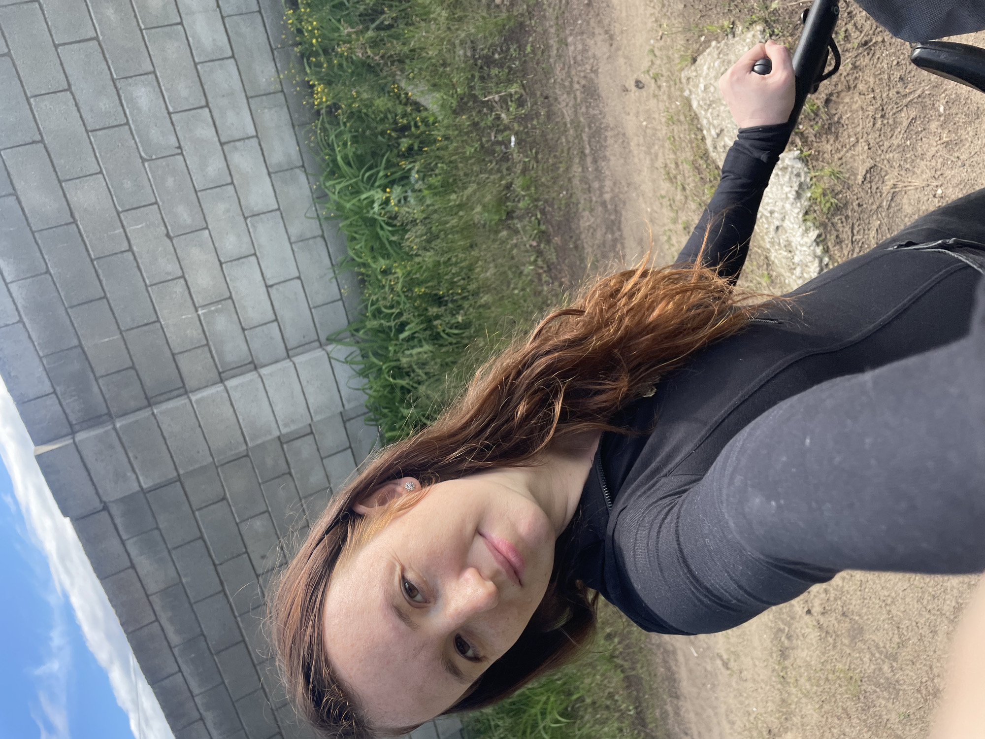 This image, captured in Vasilevka, Russia, features a female subject taking a selfie outdoors. The subject, appearing to be in her 20s or early 30s, has long, wavy reddish-brown hair and a light complexion with visible freckles. She is wearing a black long-sleeved top with a partial zipper closure at the neck. A small, silver-toned, snowflake-shaped stud earring is present in her right earlobe. She looks directly at the camera with a slight, closed-mouth smile. Her right hand is extended towards the camera, gripping a black handle, which is part of an unseen object, possibly a bicycle or stroller, partially visible in the lower right corner. The background consists of a grey concrete block wall running diagonally from the upper left. At the base of the wall and along the foreground, there is green grass mixed with small yellow wildflowers. The ground in the foreground and midground is an unpaved dirt path. A segment of blue sky with white clouds is visible in the upper left.