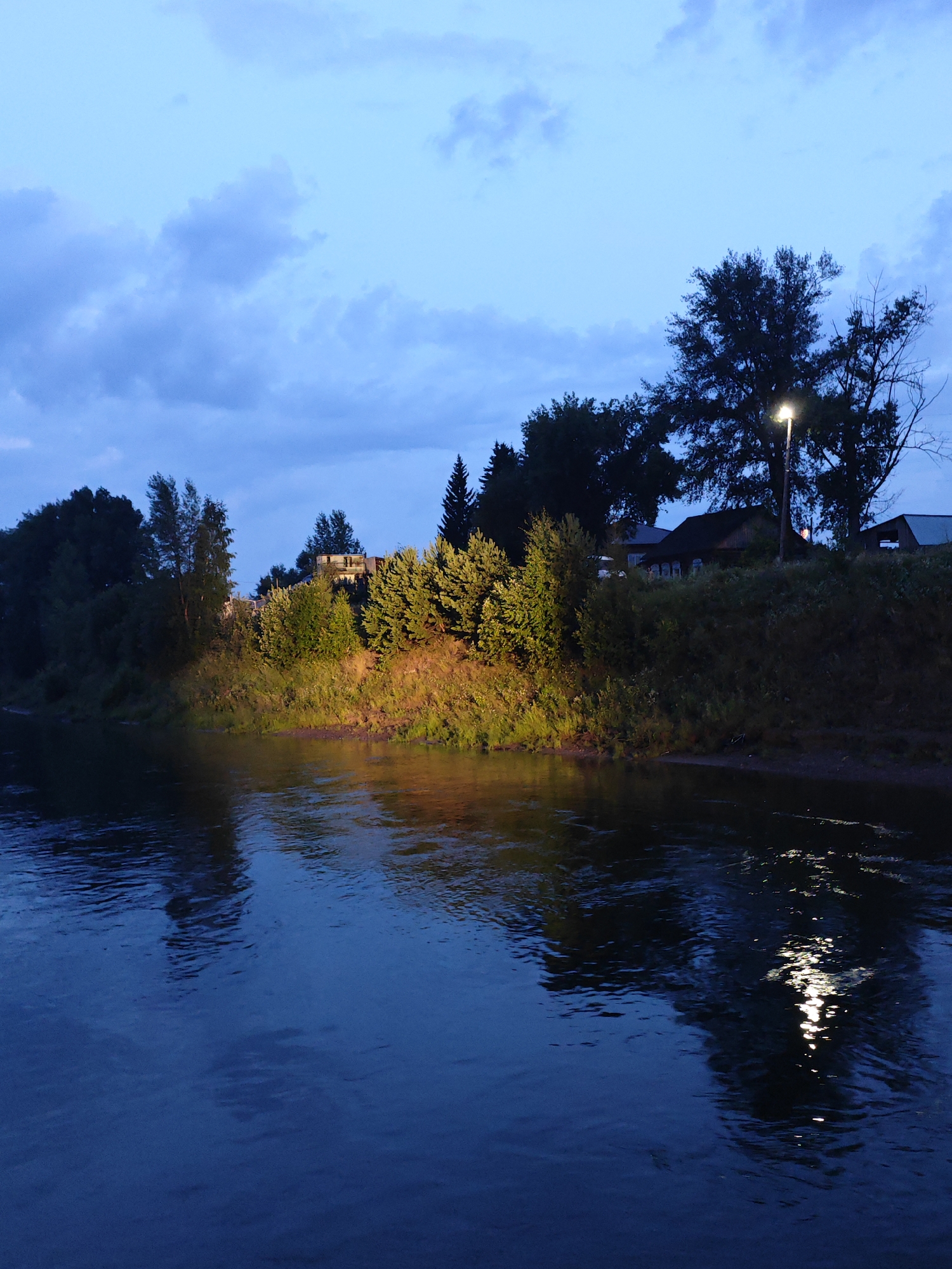 The image depicts a tranquil evening scene in Galanino, Russia, focusing on the riverbank. The sky is a deep twilight blue, with scattered clouds. On the right bank, a residential area is visible, with houses and trees. A streetlamp casts a bright light on a large tree and its reflection shimmers on the dark, calm water of the river. The vegetation on the bank is lush and green, with some areas illuminated by artificial light. The river occupies the foreground, its surface reflecting the twilight sky and the lights from the shore.