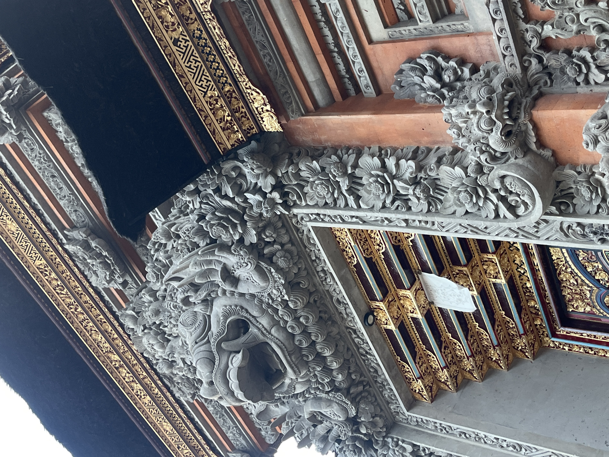 A low-angle, close-up shot depicts ornate architectural details, likely of a Balinese structure, in the Ubud District of Indonesia. Predominantly constructed of gray stone, the scene features intricate carvings of floral patterns and grotesque figures with open mouths and prominent teeth. Accents of gold filigree and red and blue paint are interspersed amongst the stone, enriching the visual complexity. The perspective leads the eye upward, emphasizing the height and artistry of the architectural element.