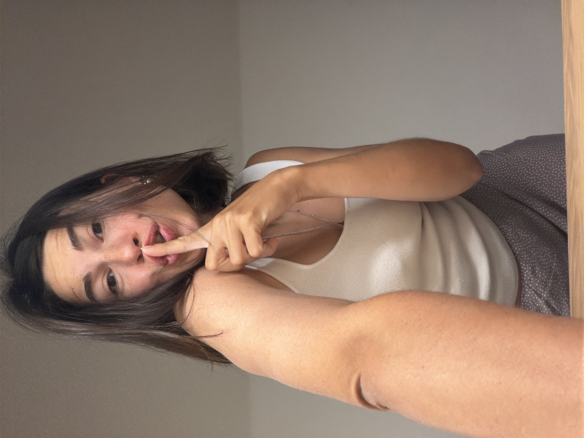 A young woman with dark brown hair is looking at the camera. She is wearing a white tank top and is holding her finger to her lips. Behind her, there is a light gray wall and a brown patterned fabric. There is no indication of the location being Budva, Montenegro.