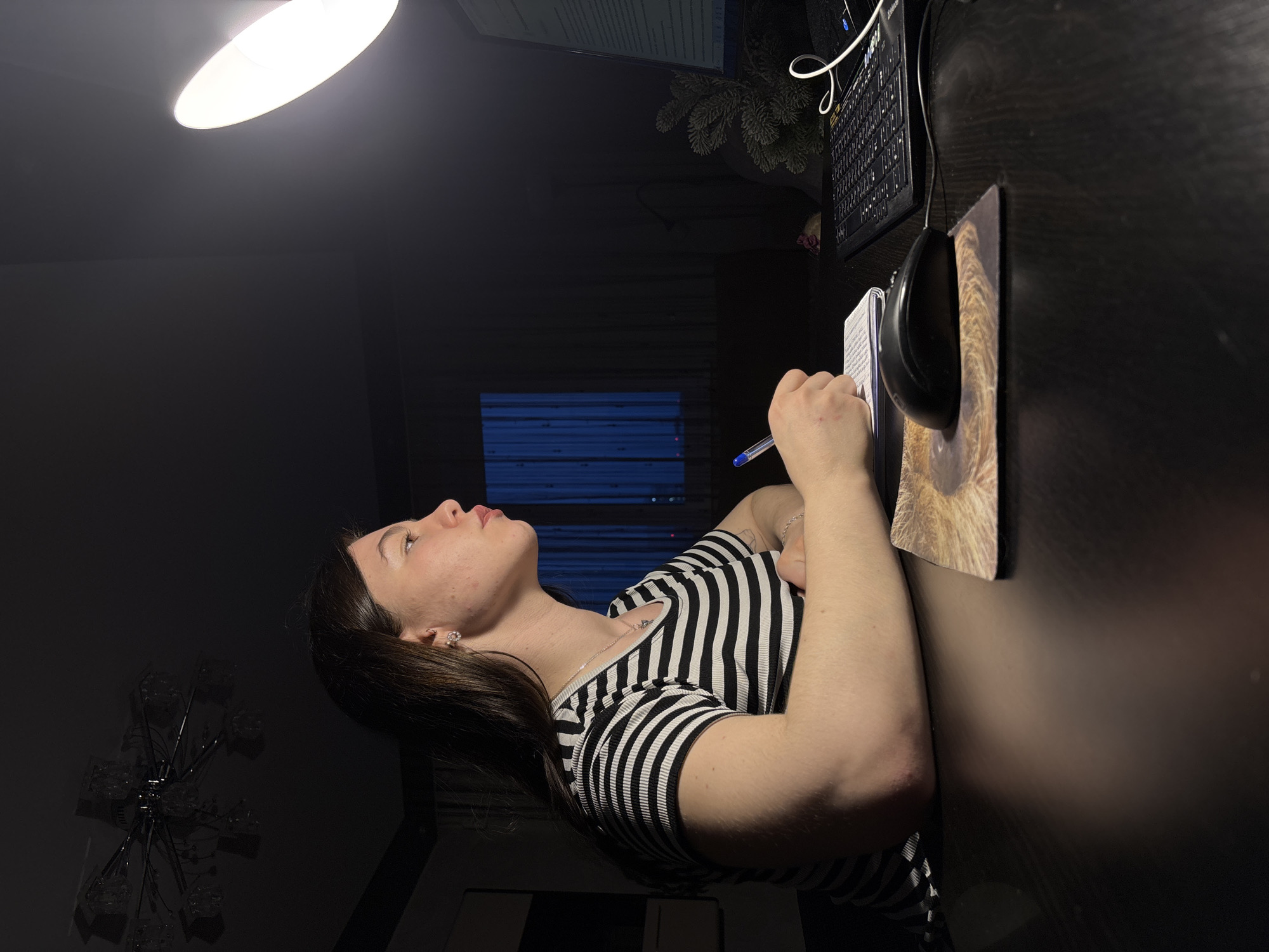 The image depicts a person, presumably female, with long dark hair and a striped black and white shirt, sitting at a dark-colored desk. She is looking upwards and holding a pen in her right hand, positioned near a notebook. On the desk, there's a computer keyboard, mouse, and a mousepad with a design featuring a lion's eye. A desk lamp illuminates the scene from above. The background is dark, with a window visible, and the reflection of a chandelier-like light fixture can be seen on the desk surface. The location is assumed to be in Perm, Russia, based on the image context.
