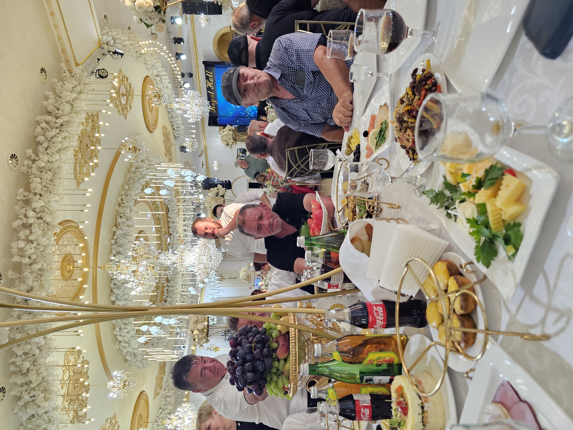 The image captures a formal indoor event, likely a banquet or wedding reception, taking place in the "Royal Hall" in Abazinka, Russia. The opulent ceiling is adorned with elaborate gold trim, numerous crystal chandeliers, and cascading white floral arrangements, with additional delicate white flowers and string lights decorating the walls. Attendees are seated at a long, white-clothed banquet table laden with food and drinks. In the foreground, a tiered fruit display with grapes, apples, and other fruits sits prominently. Various bottles of Coca-Cola and other beverages are visible, along with plates of appetizers, including what appears to be small pastries with melted cheese, a layered salad, and platters of cheese and smoked salmon. Several men are the primary focus, dressed in casual and semi-formal attire. One man in a blue checkered shirt and a baseball cap is looking to the right, with a slight smile. Another man in a white polo shirt is reaching towards the fruit display, while a man with a beard in a black t-shirt is looking down at the table. The overall atmosphere suggests a celebratory gathering with a luxurious setting.